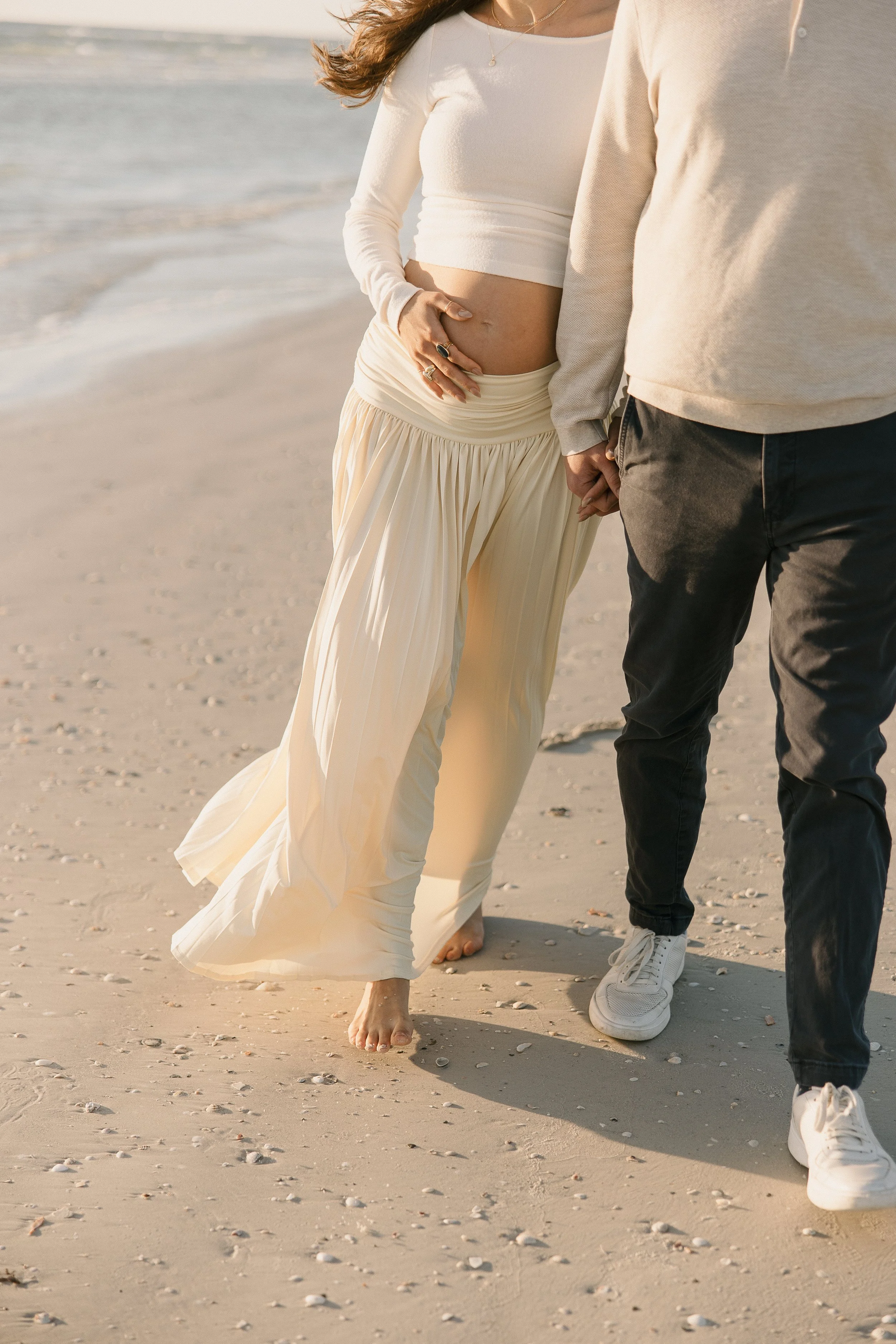 A pregnant woman in a white crop top and flowing skirt walking barefoot on a sandy beach, holding hands with a person wearing dark pants and white sneakers, with the ocean in the background.
