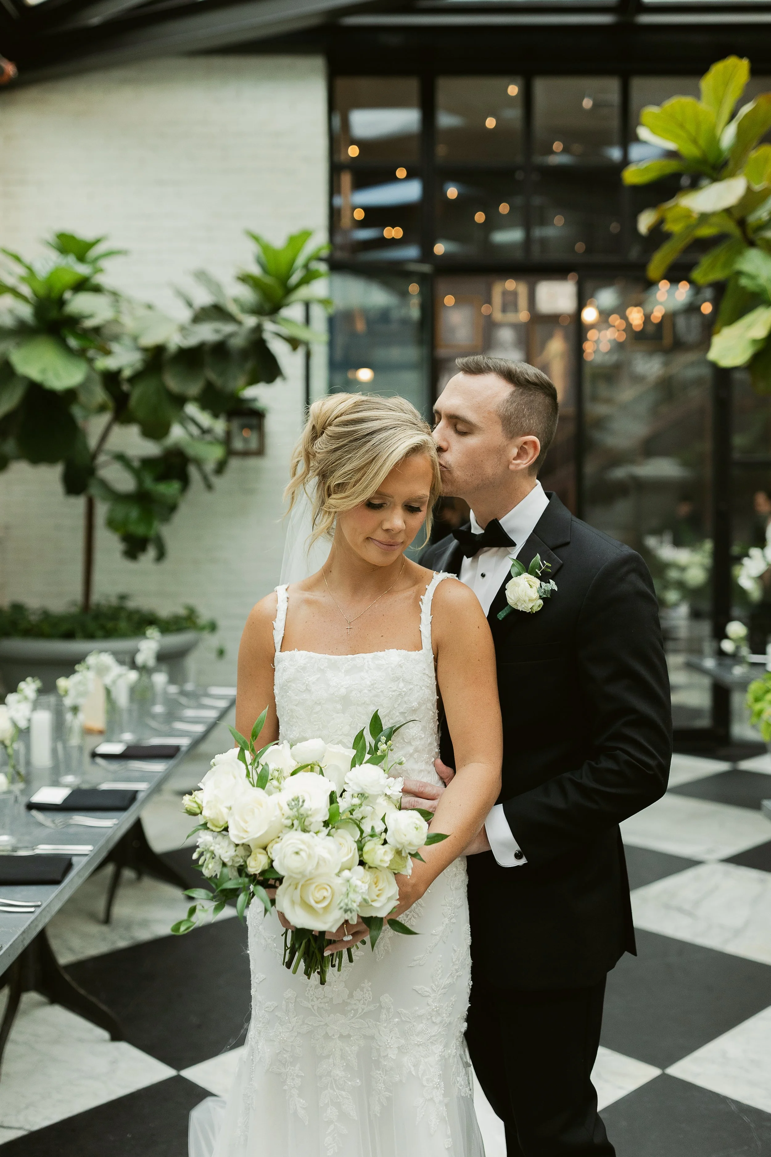 A newlywed couple standing close together during their wedding celebration. The bride has blonde hair styled in an updo with a white veil and is holding a bouquet of white flowers. The groom, wearing a black tuxedo with a white shirt and black bow ti