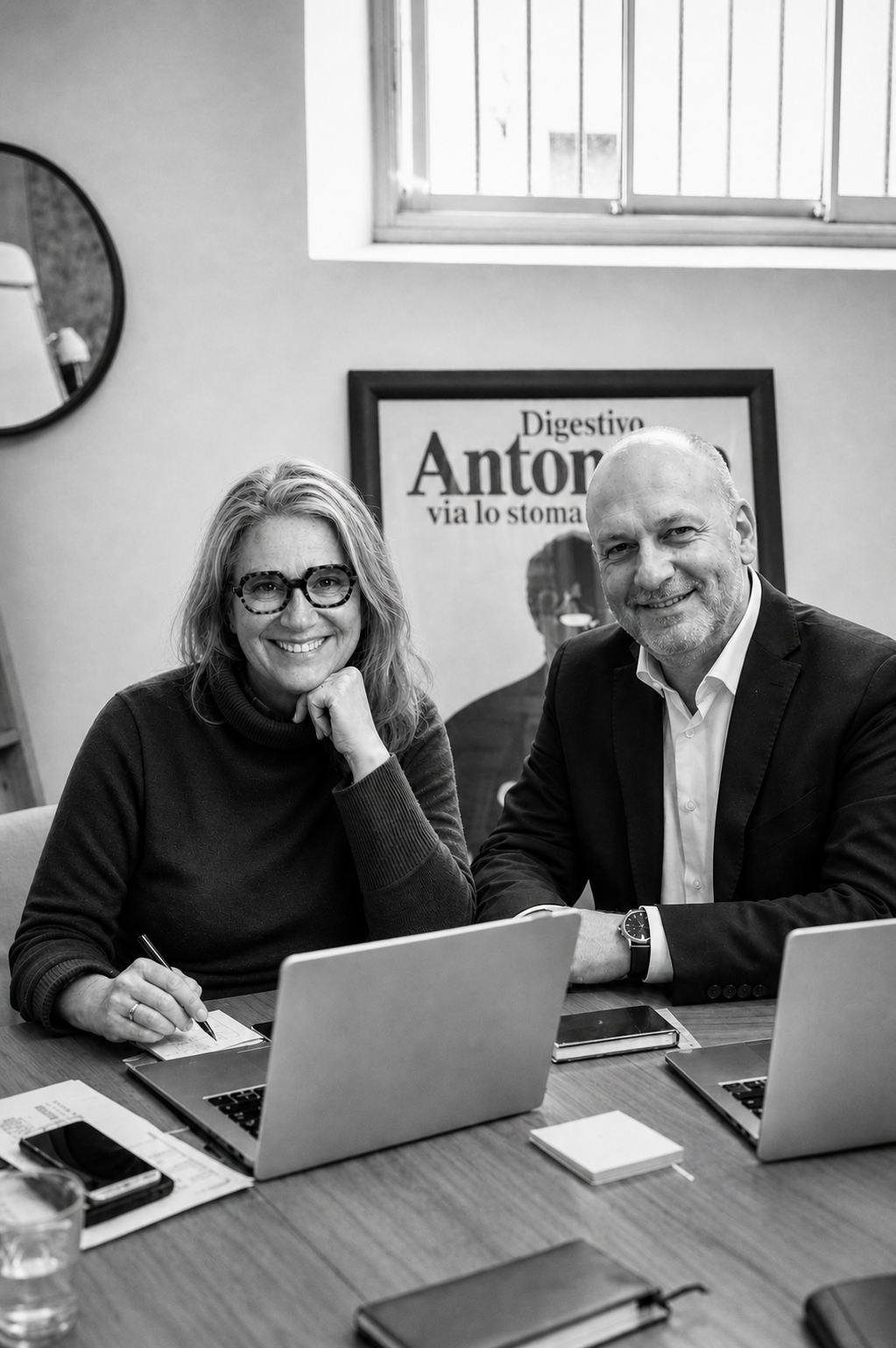 Deux personnes, un homme et une femme, assises à une table de bureau, souriant à la caméra, avec des ordinateurs portables devant eux, dans un bureau avec une affiche en arrière-plan.