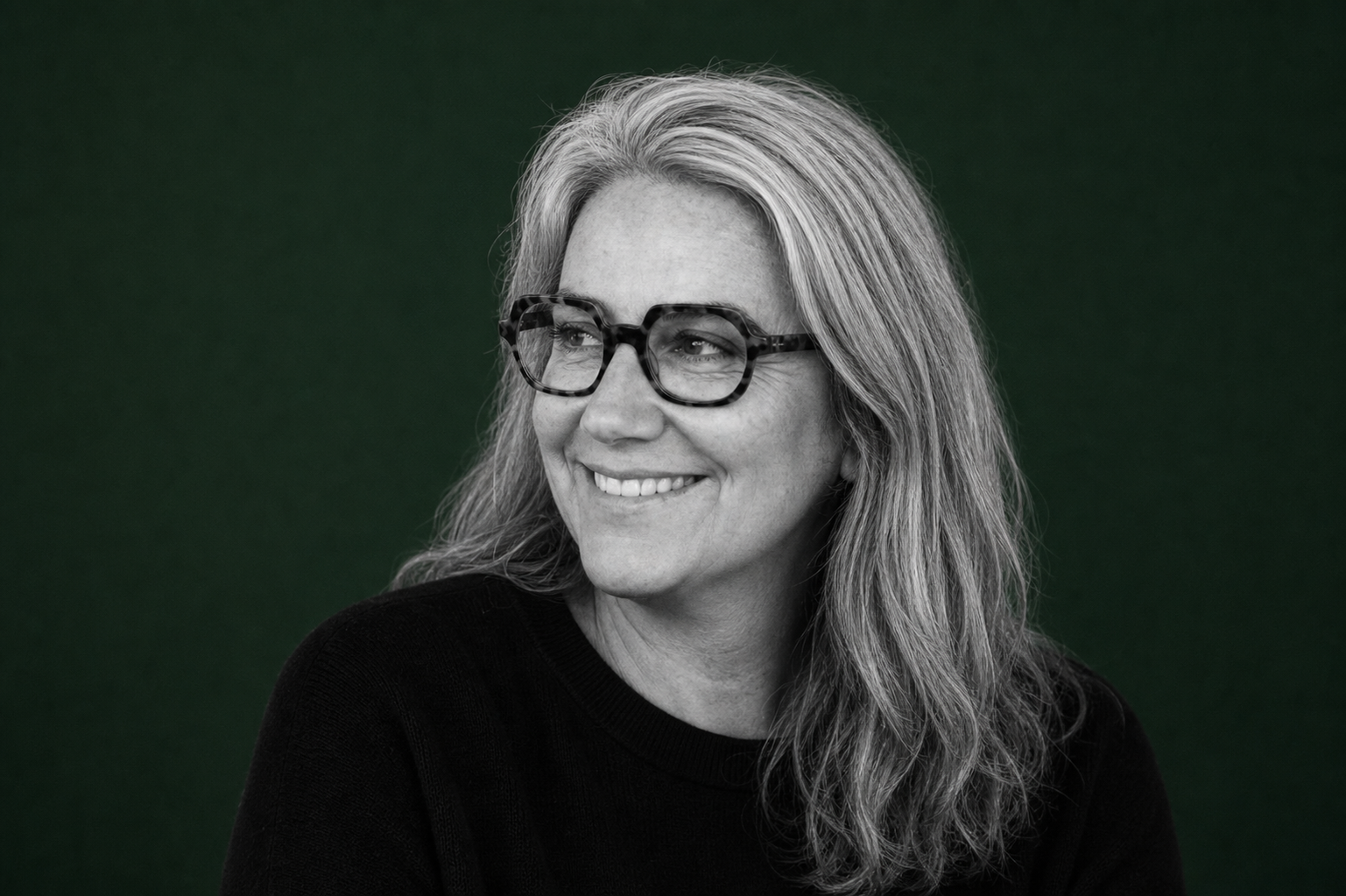 Une femme portant des lunettes, souriante, avec des cheveux longs et gris, vêtue d'un pull noir, contre un fond vert.