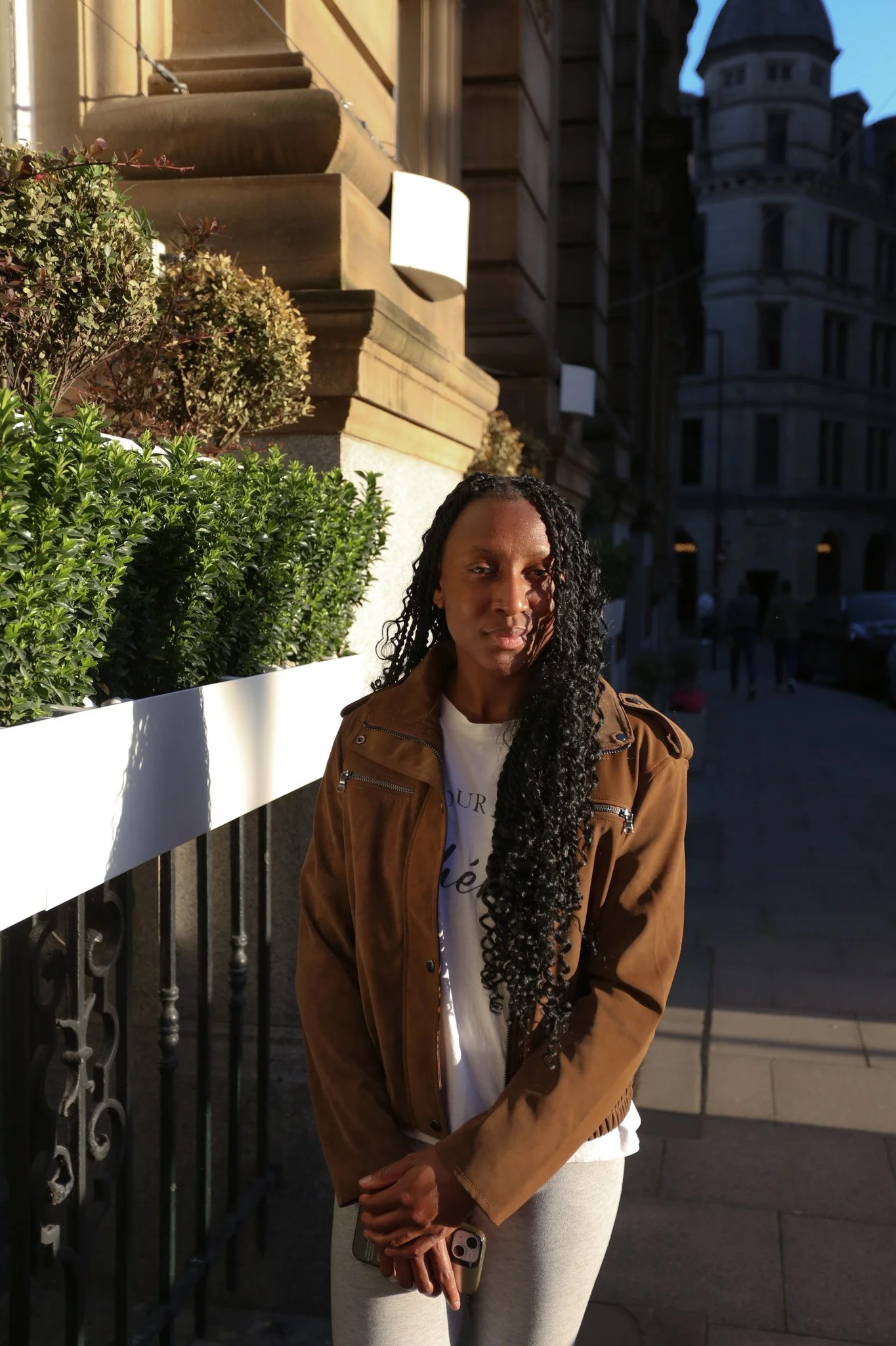 A woman with long, curly black hair in a ponytail stands on a city street in front of a building with ornate architecture. She is wearing a brown jacket, light-colored pants, and holds a smartphone in her right hand. The background shows a dark build