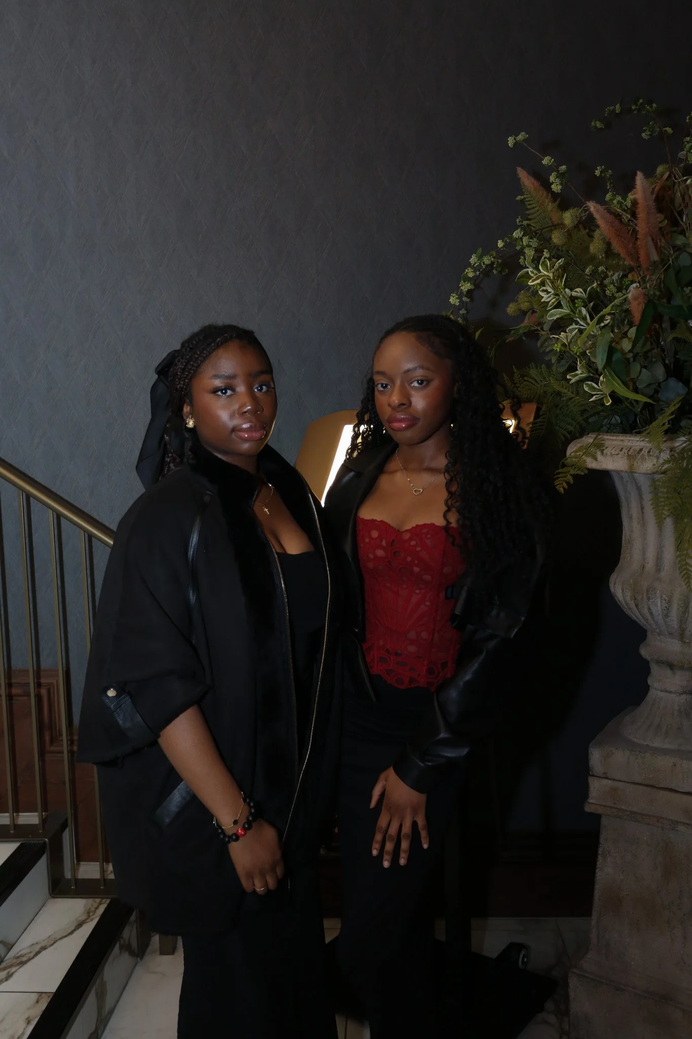 Two young women standing together indoors, dressed in formal attire, with dark hair, one wearing a black jacket and the other in a red lace top, near a large decorative plant and a stone column.
