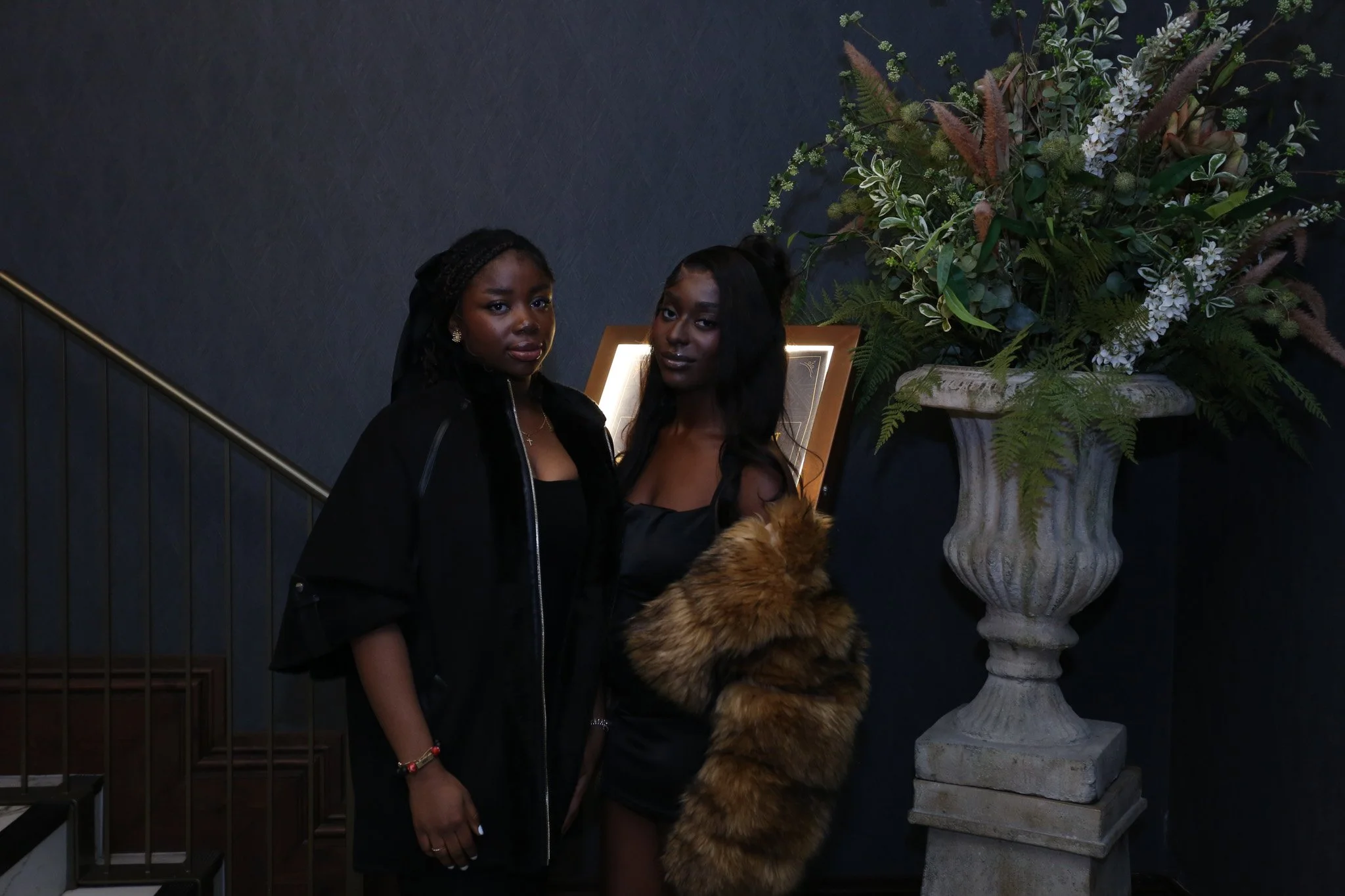 Two women standing beside a large floral arrangement with greenery, white flowers, and ferns in an ornate stone vase, indoors against a dark wall.