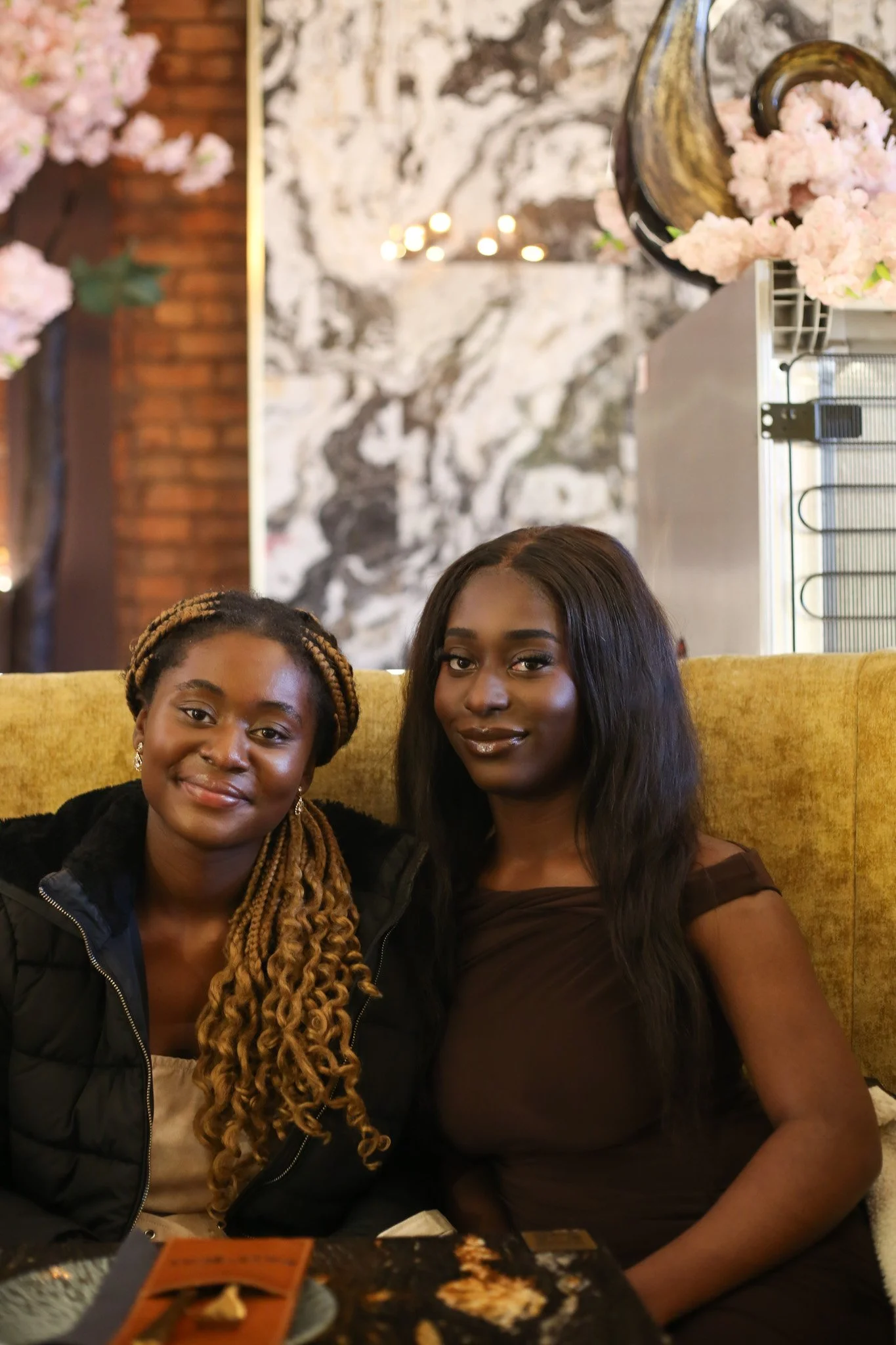 Two women sitting close together in a restaurant, smiling, with a marble wall and flowers in the background.