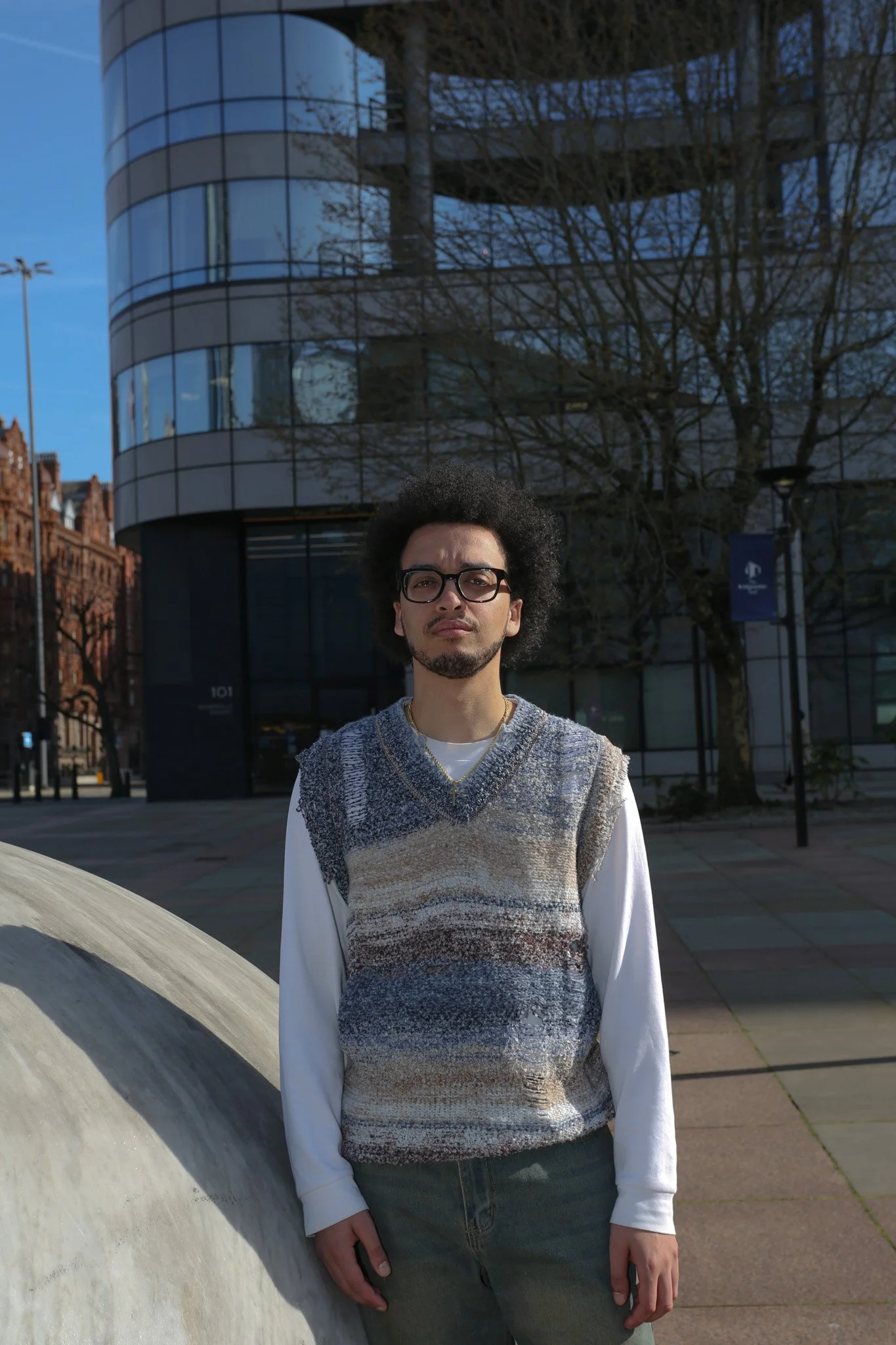 A man with glasses and curly hair standing outdoors in front of a modern glass building with reflections of trees and a blue sky. He is wearing a multicolored sweater over a white shirt and gray jeans.