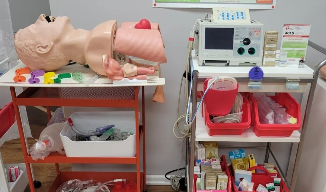 Medical training setup with a CPR dummy head, lung and airway models, and various medical supplies, including a defibrillator, medications, and medical instruments on tables.