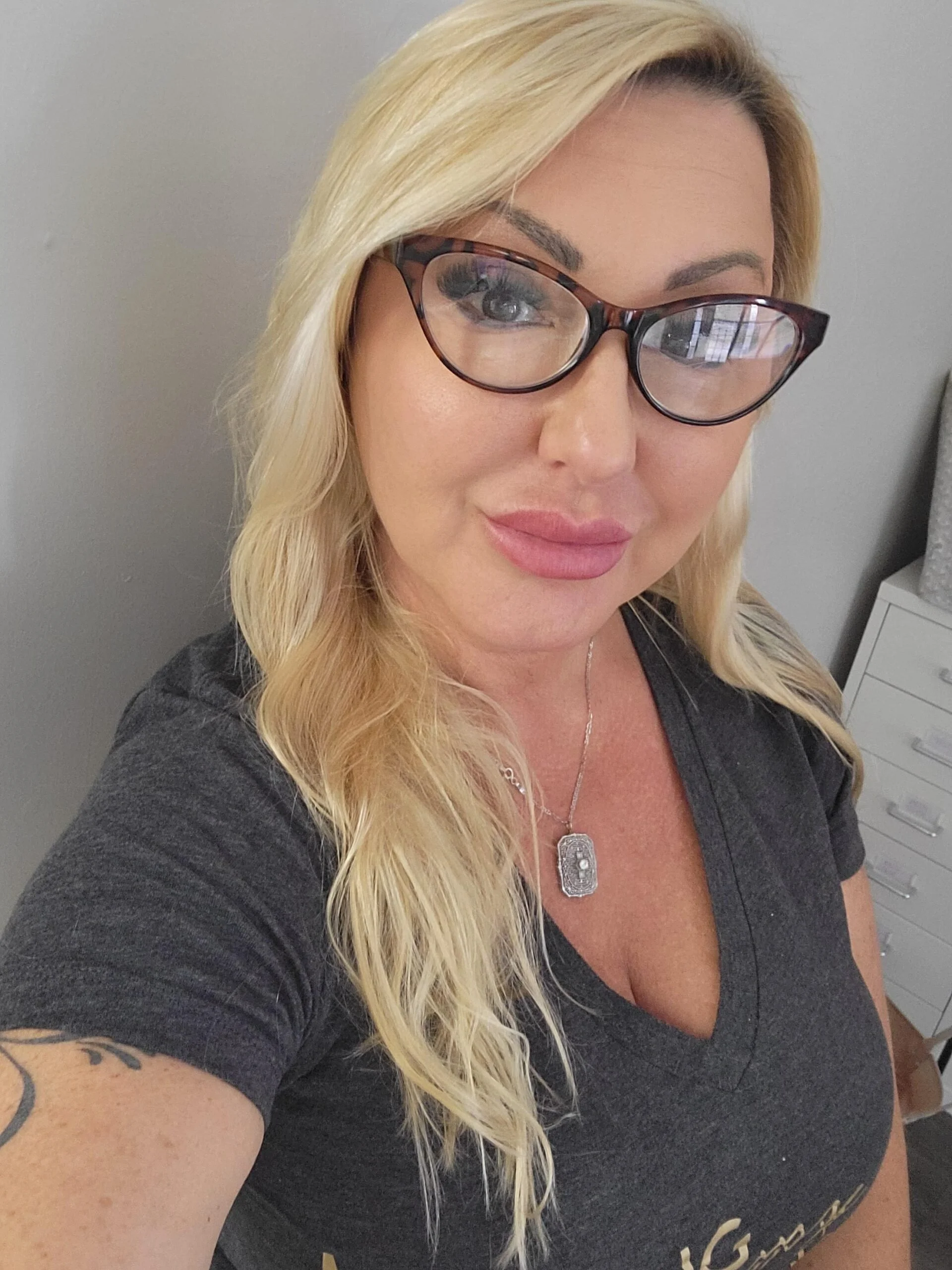A woman with long blonde hair, wearing glasses with a tortoise shell pattern, a black shirt, and a silver necklace with a rectangular pendant. She is taking a selfie in a room with a gray wall and a white dresser with drawers in the background.