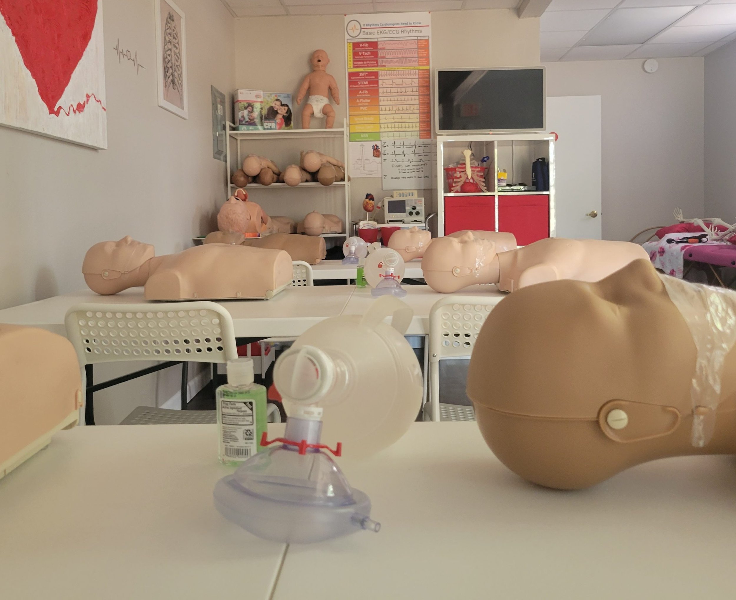 Medical training room with CPR manikins on tables, a poster of ECG rhythms on the wall, and shelves with additional manikins and medical supplies.