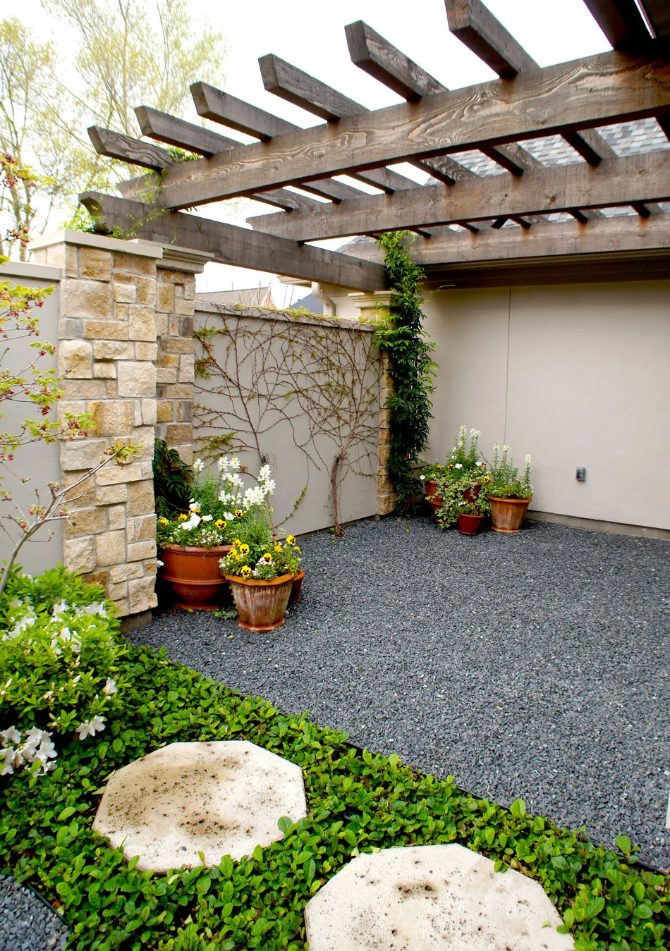Wood stained pergola offers shade to well appointed outdoor seating area. White azaleas and container planted seasonal color give this secret garden a playful appeal.