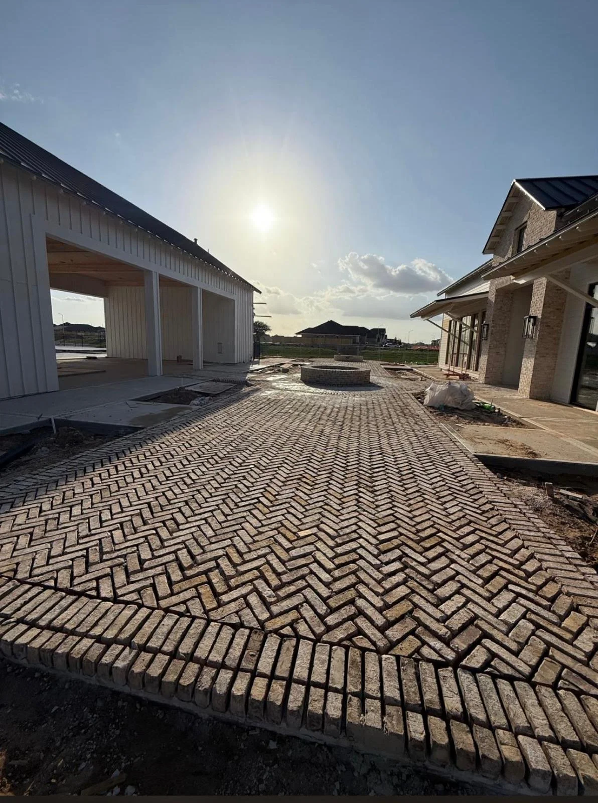 A construction site with brick paver driveway in progress, modern houses under construction on the right and left, with a clear sky and the sun shining.