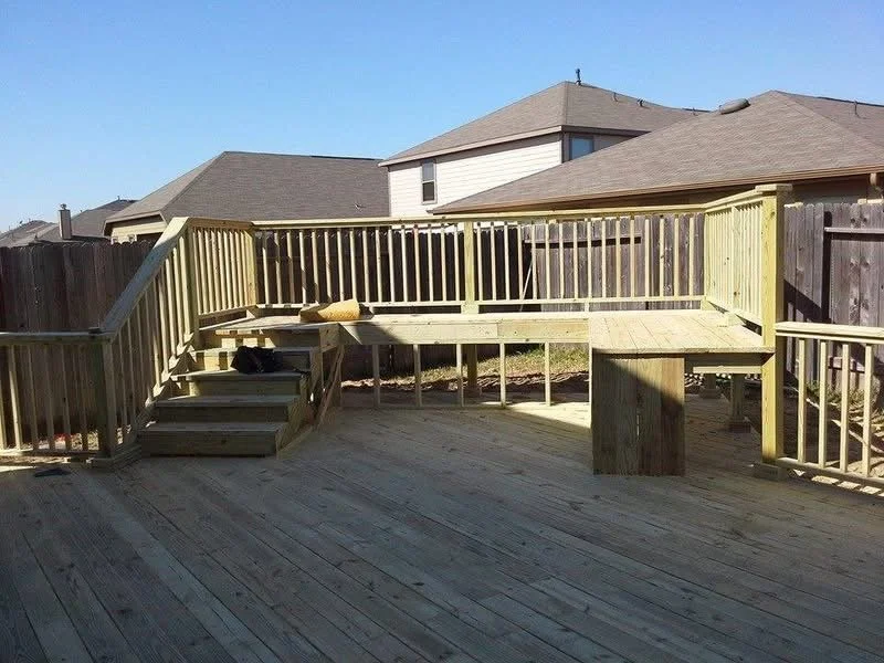 Newly built wooden deck with stairs and safety railing in a backyard, surrounded by neighboring houses under a clear blue sky.
