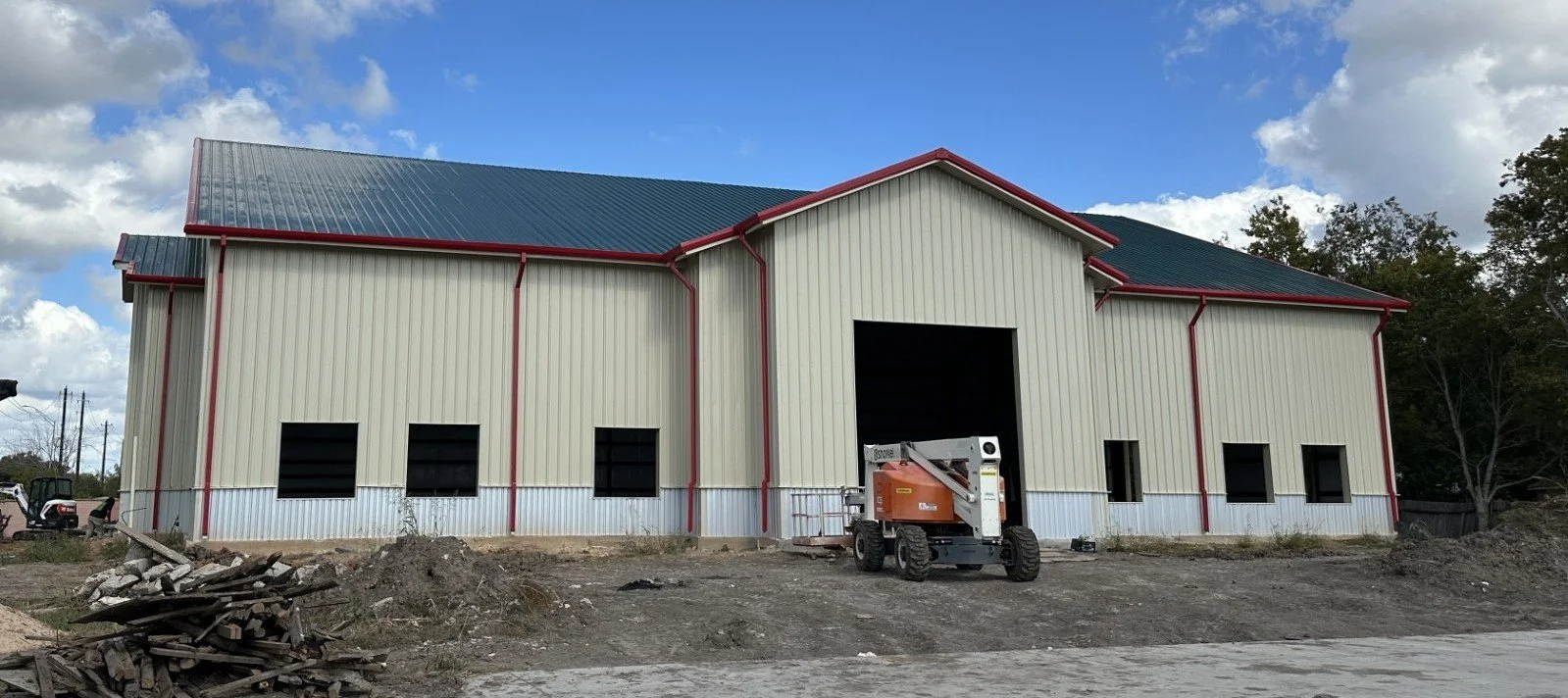 An industrial building under construction with beige metal siding, red trim, and a blue metal roof. There is construction equipment in front and trees in the background.
