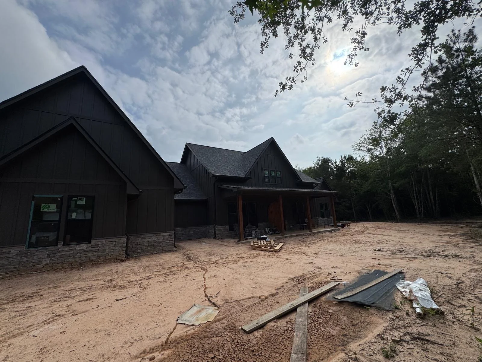 A house under construction with black siding, stone accents, and a covered porch, surrounded by dirt and construction materials, with trees in the background and a cloudy sky above.