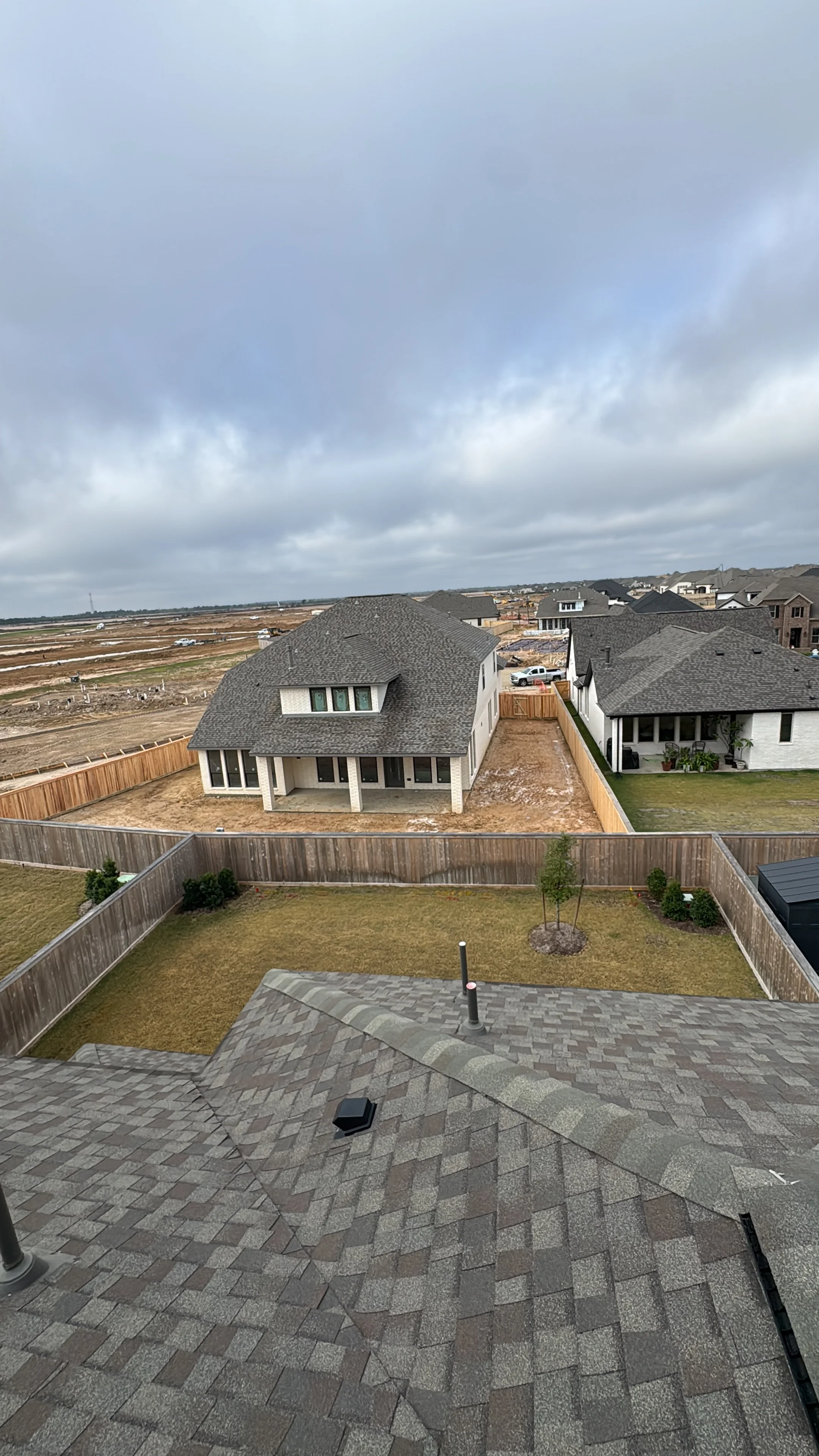 View from a house's roof showing a backyard with grass, trees, and a wooden fence, neighboring houses, and an overcast sky.