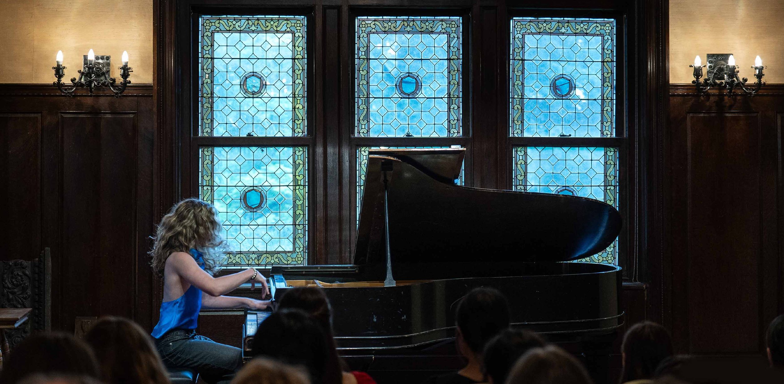 Young girl playing grand piano in a room with stained glass windows and wall-mounted lamps