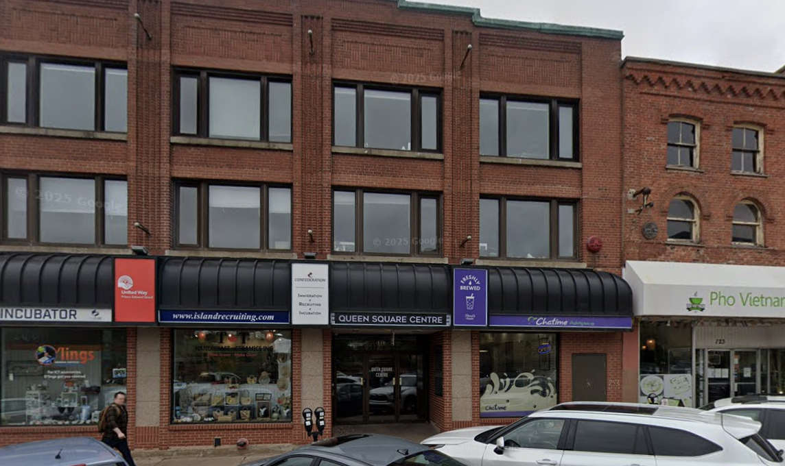 A brick building with multiple business signs on the ground floor, including a recruitment agency, a café named  Chatime,  and a pharmacy called  Pho Vietnam. Cars are parked in front of the building, and a person is walking on the sidewalk.