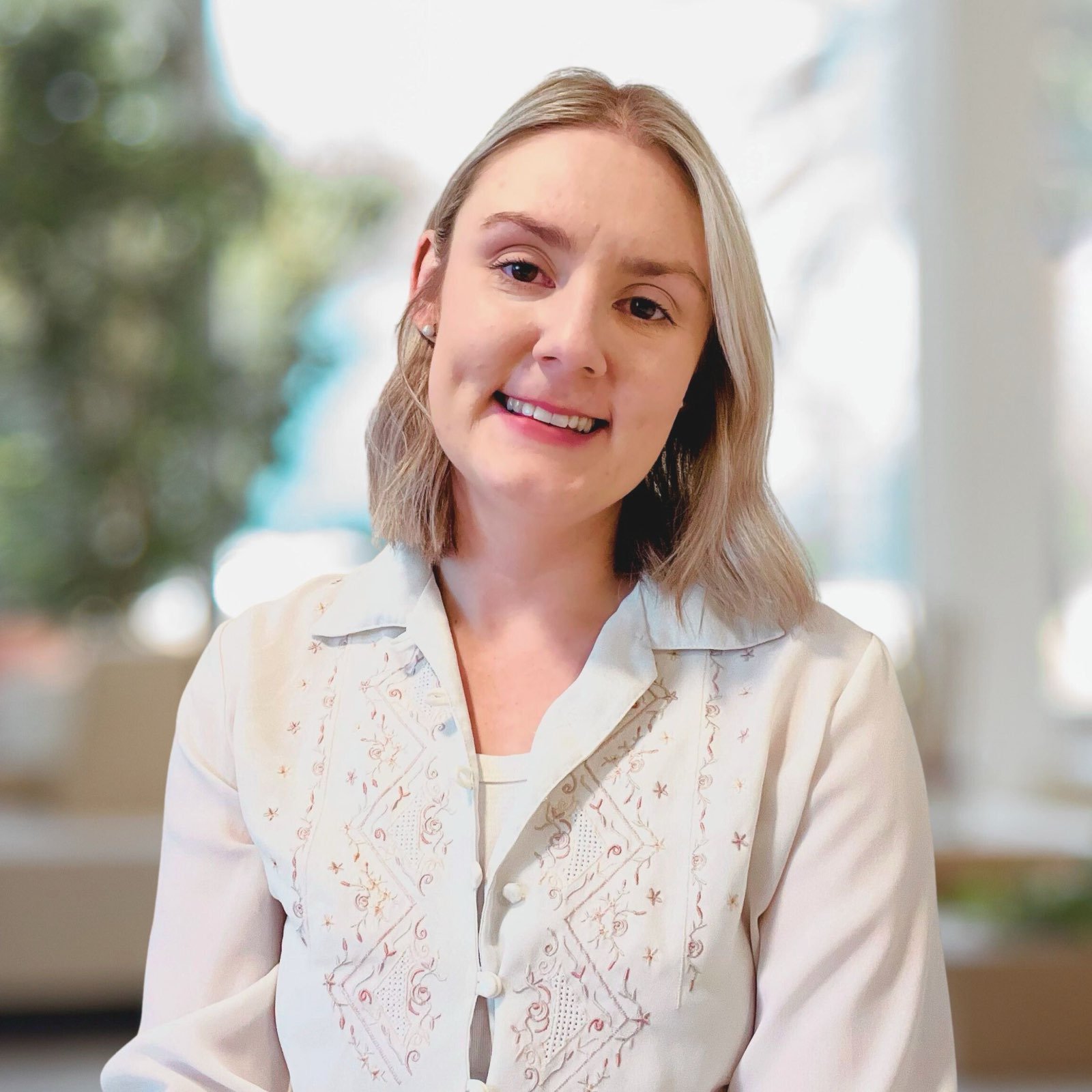 A woman with blonde hair smiling at the camera, wearing a white embroidered blouse, standing outdoors with sunlit blurry trees in the background.