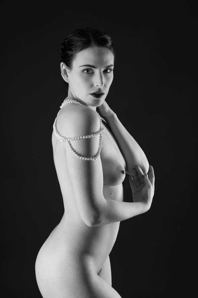 Black-and-white fine-art photograph of a nude woman adorned with pearl jewellery, standing against a dark background and looking towards the camera.
