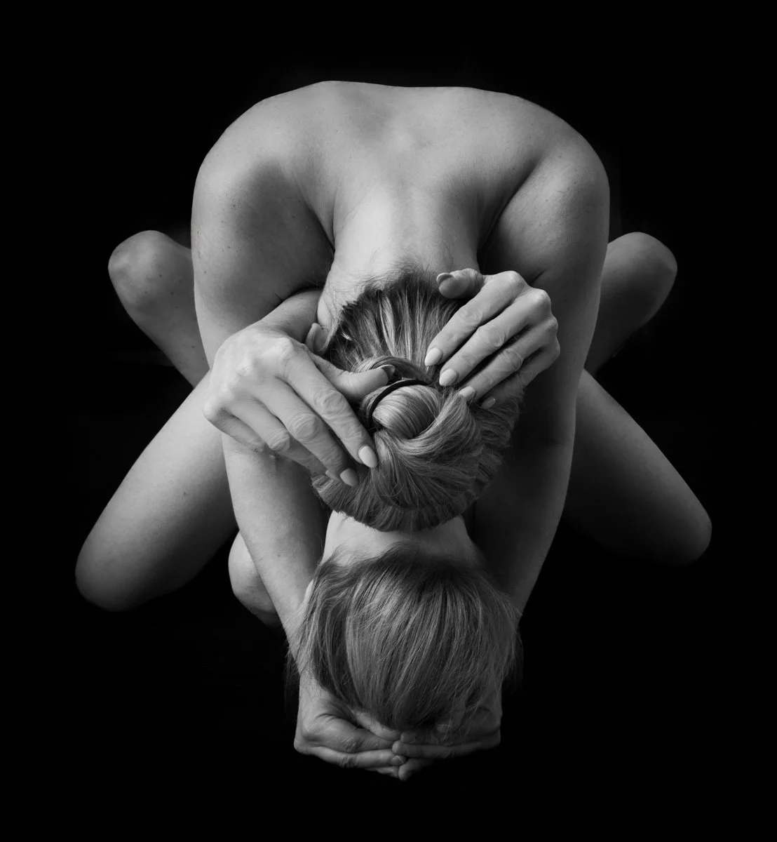Black-and-white fine-art photograph of two intertwined nude female figures, photographed from above against a dark background.