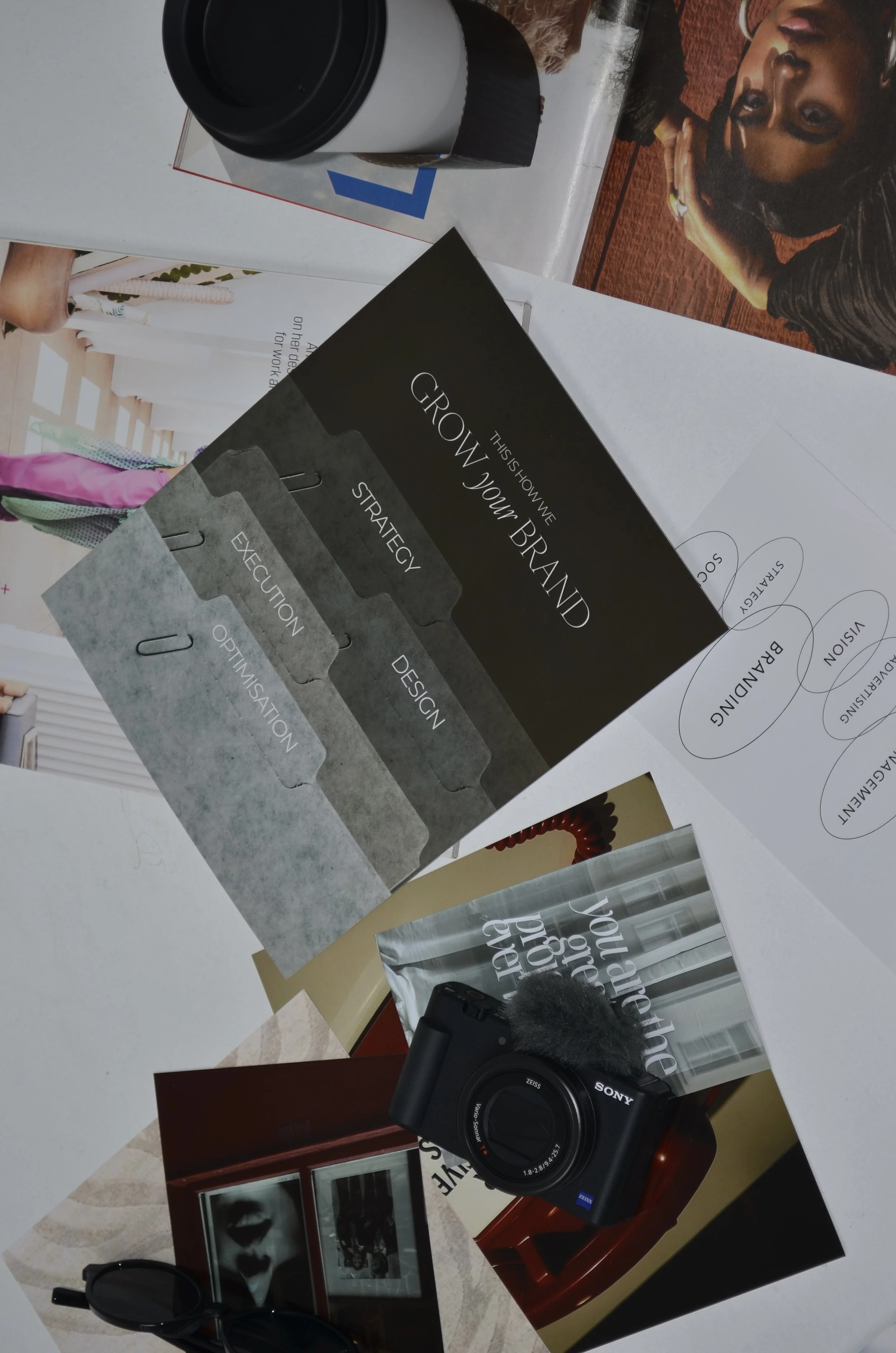 A desk with various fashion and branding materials, including a book titled 'Grow Your Brand' with sections on strategy, execution, and branding, a camera, sunglasses, photographs, and a coffee cup.