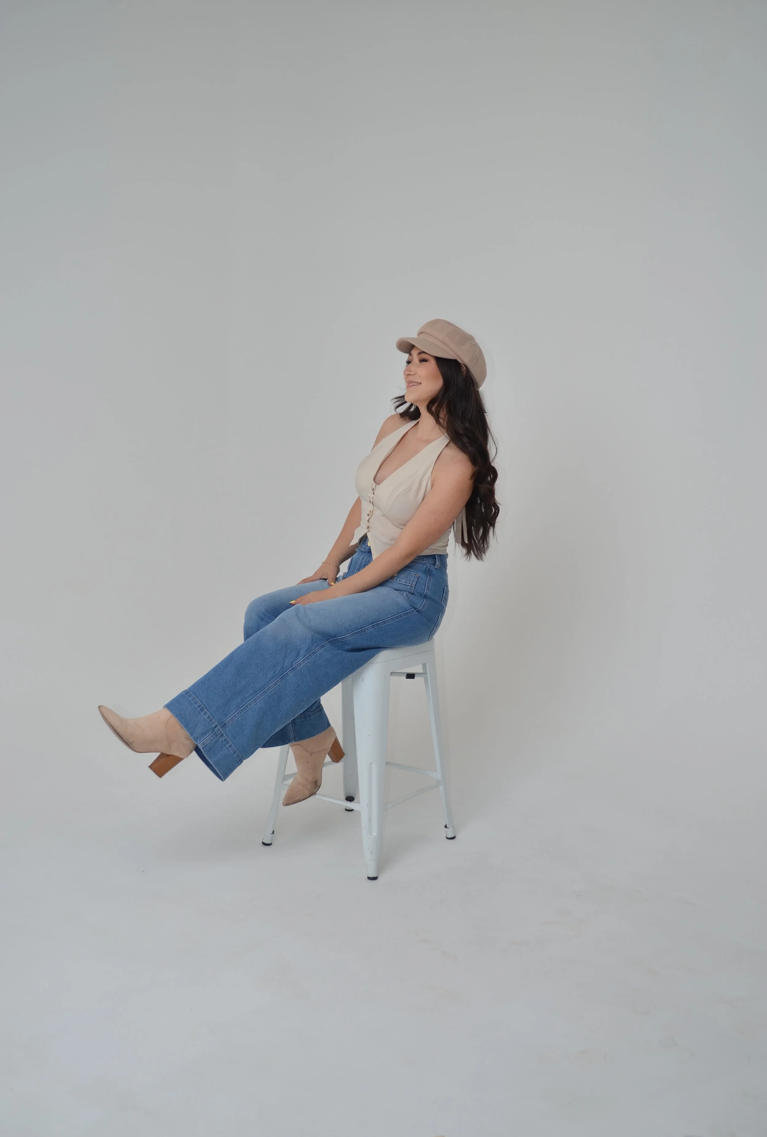 A woman sitting on a white stool, wearing a beige hat, cream sleeveless top, high-waisted blue jeans, and beige high heels, smiling against a plain white background.