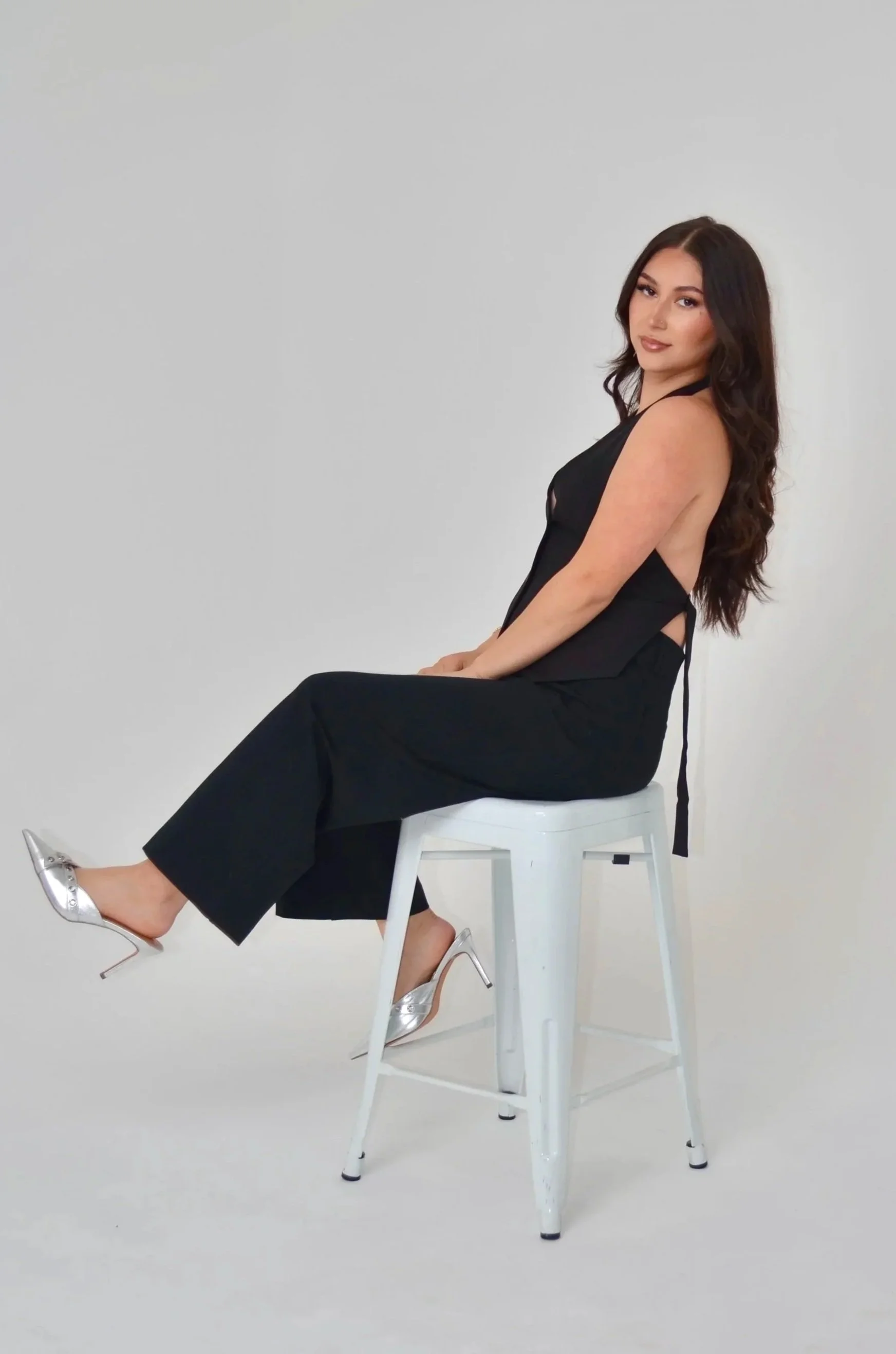 A woman with long dark hair sitting sideways on a white chair in a black sleeveless outfit and metallic silver heels, against a plain light gray background.