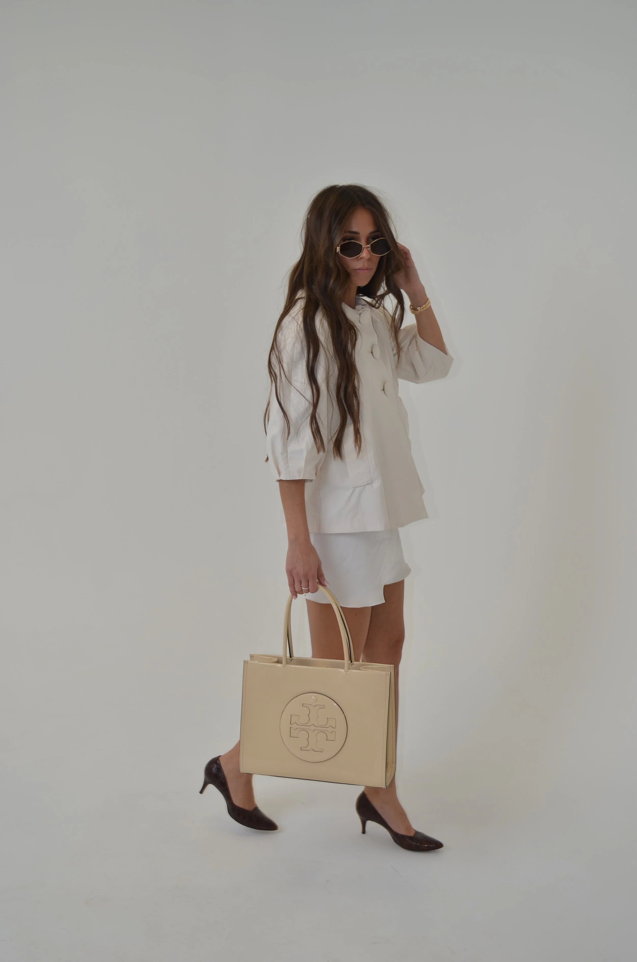 Stylish woman with long wavy brown hair wearing black sunglasses and a cream-colored blazer, holding a beige designer handbag, standing in front of a plain white background.