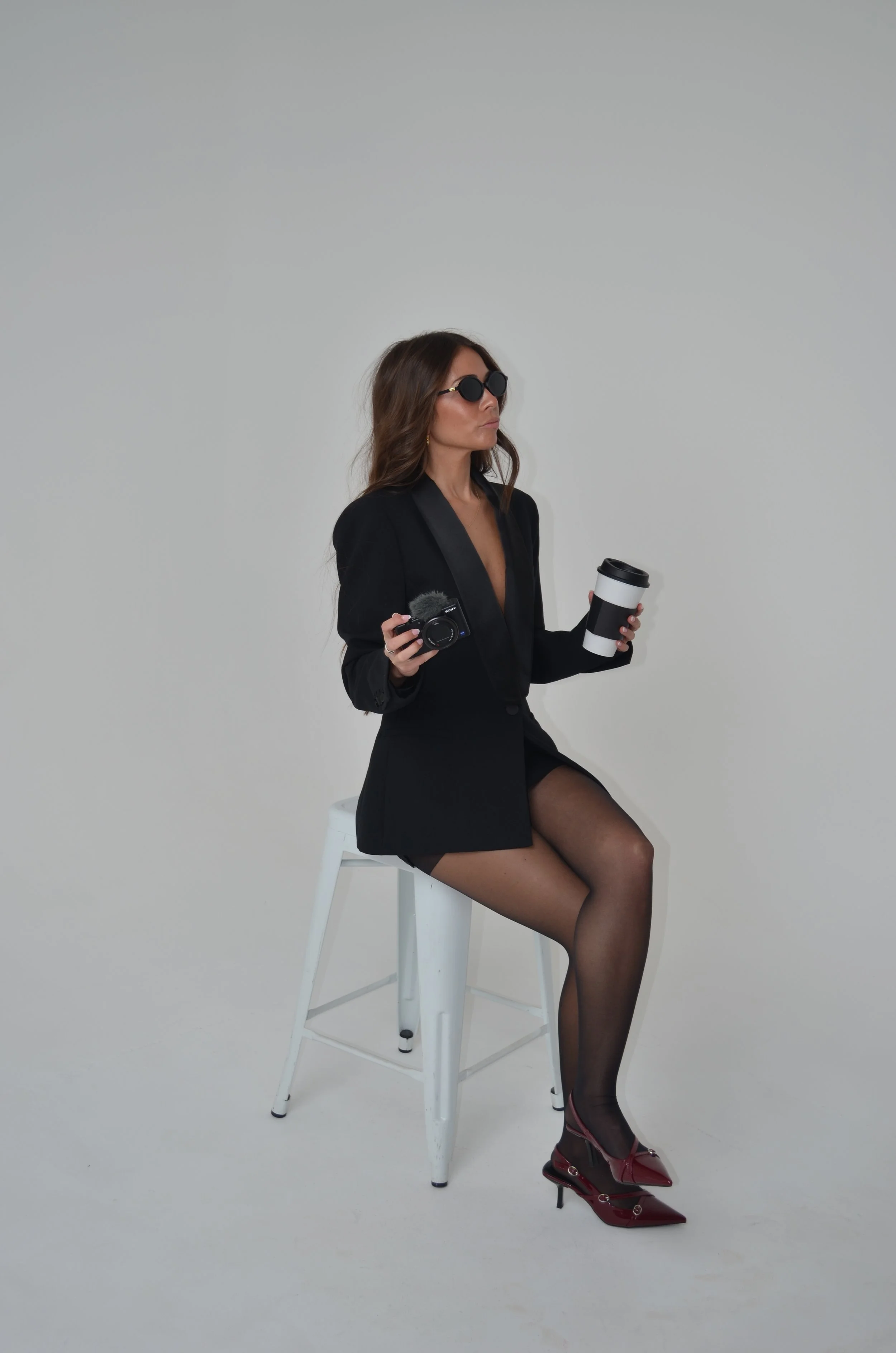 A woman sitting on a white stool against a plain light gray background. She is wearing a black blazer dress, sheer black stockings, and red high-heeled shoes. She has long brown wavy hair, black sunglasses, and is holding a travel coffee cup in one h