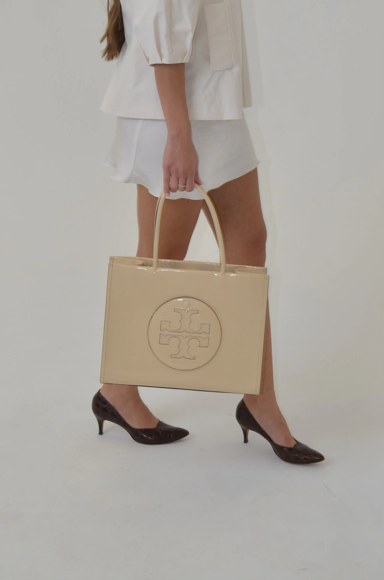 A woman dressed in a white blazer and white skirt, wearing high heels, holding a beige designer tote bag with a circular logo.