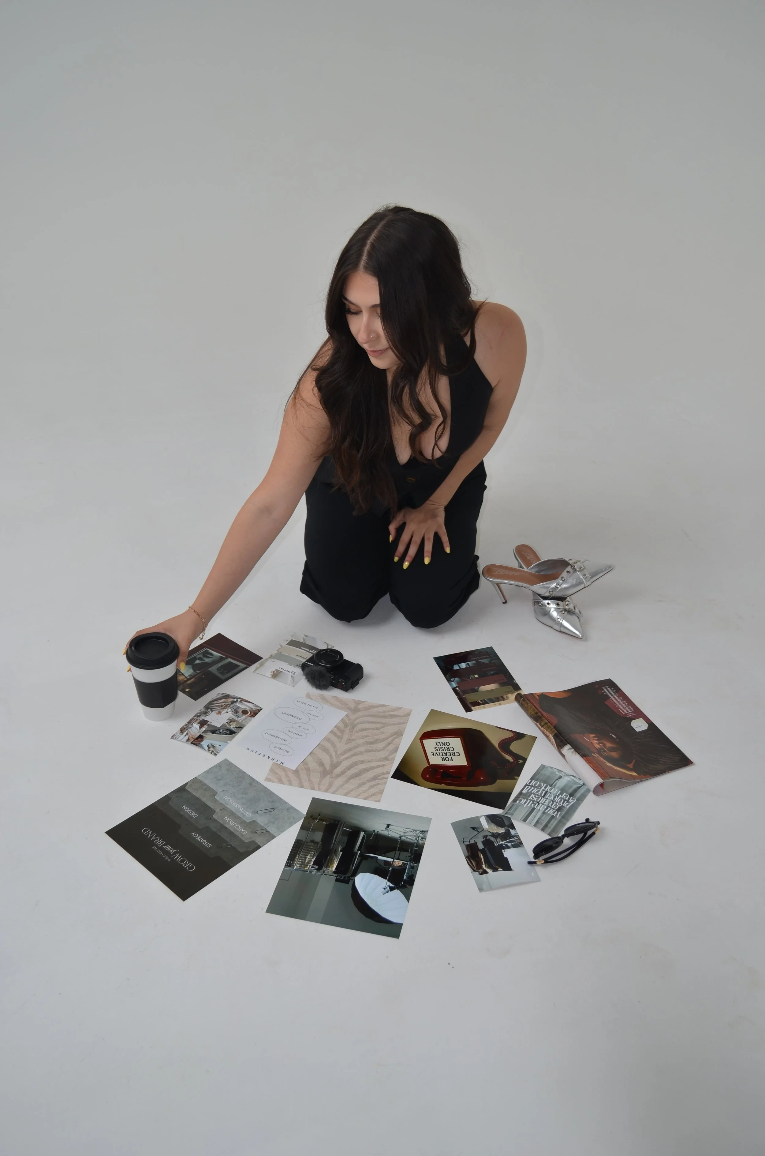 A woman with long dark hair, wearing a black top, is kneeling on a white floor, surrounded by various magazines and a camera. She holds a coffee cup and appears to be looking at or arranging the magazines.