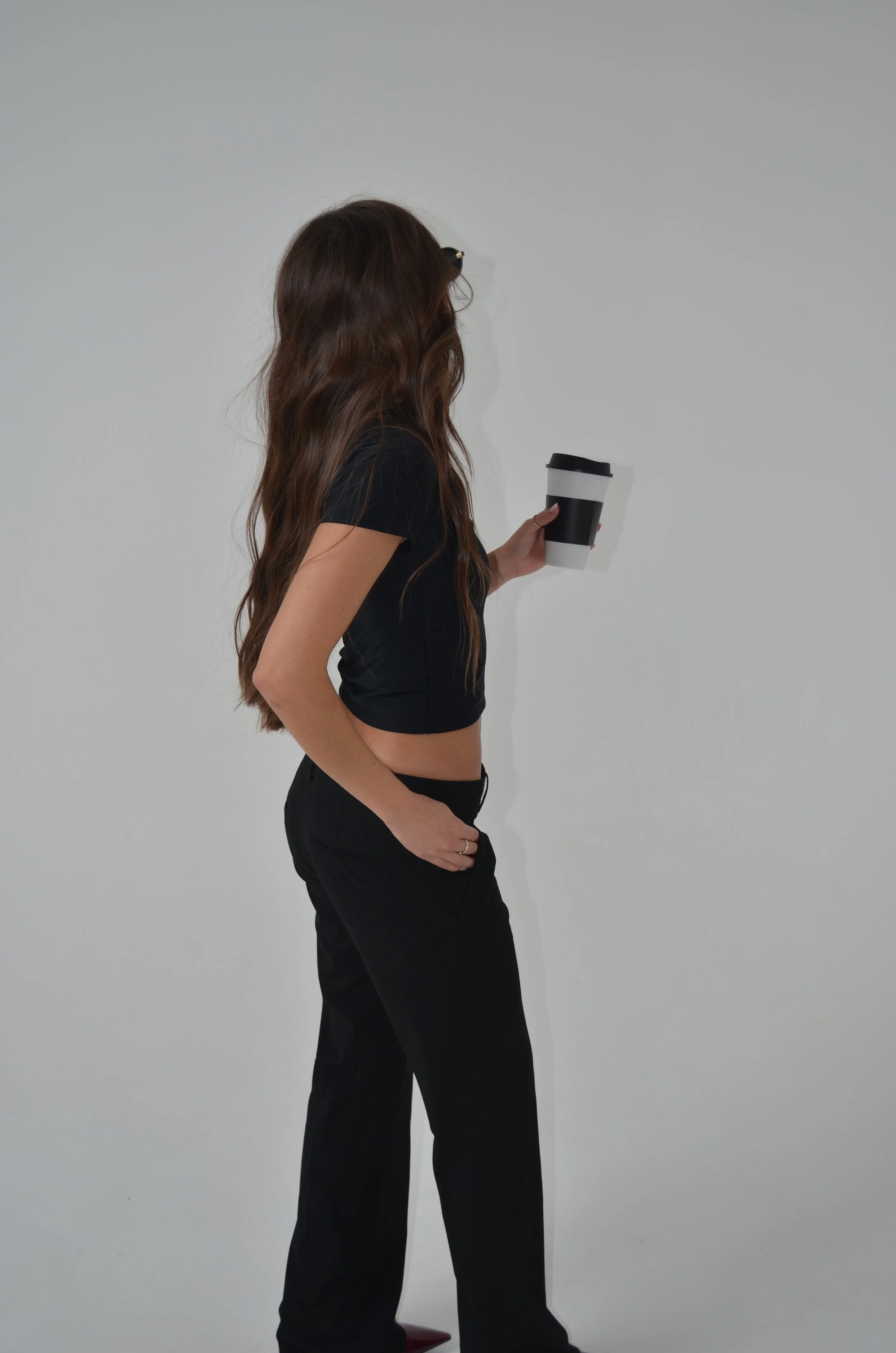 A woman with long wavy brown hair wearing black clothing holding a white to-go coffee cup with a black sleeve and black lid, standing against a plain white wall.