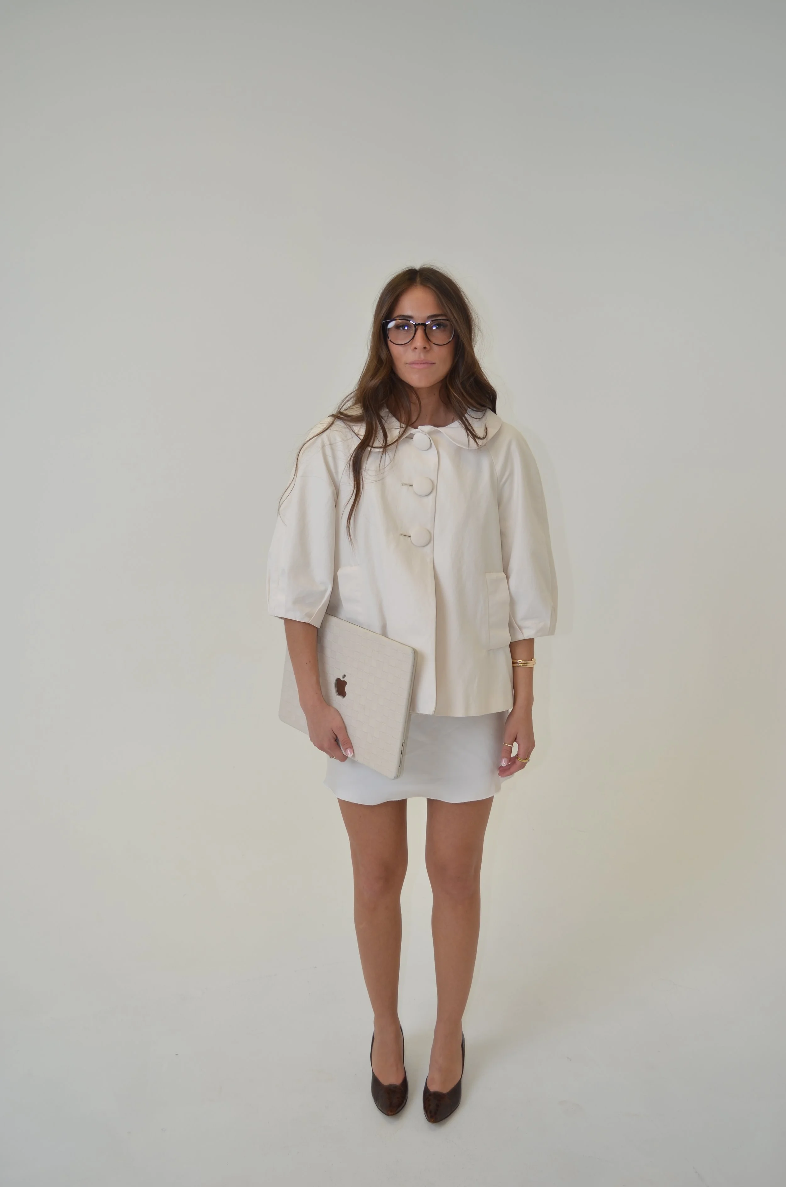 A woman standing in front of a plain white background, wearing glasses, a cream-colored jacket with large buttons, a matching skirt, and heels, holding a laptop.