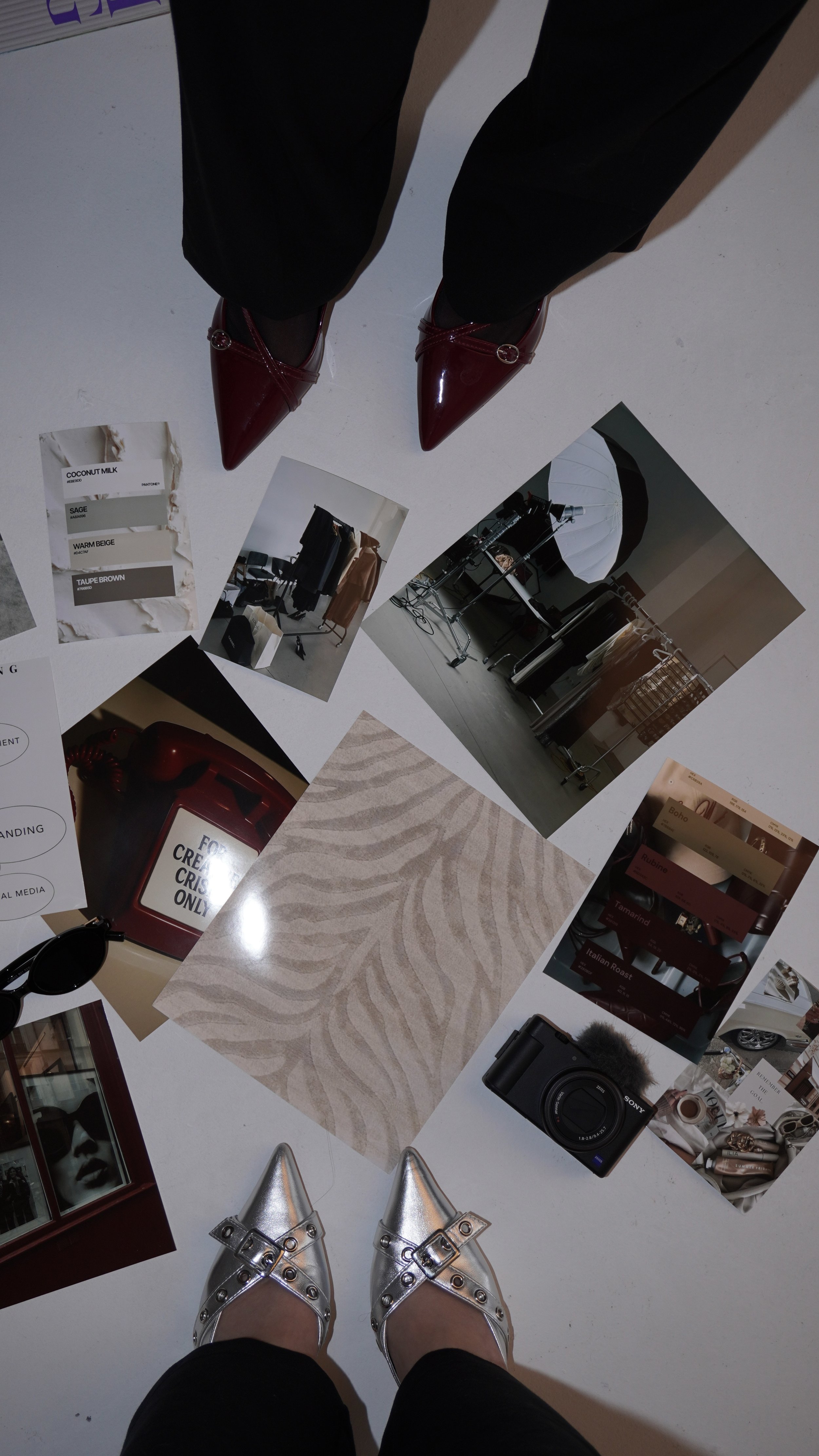 Photo taken from the person's perspective looking down at their shiny silver pointed-toe shoes with buckles, standing on a white surface scattered with various photos, swatches, and objects including a camera, lipstick samples, and clothing racks.