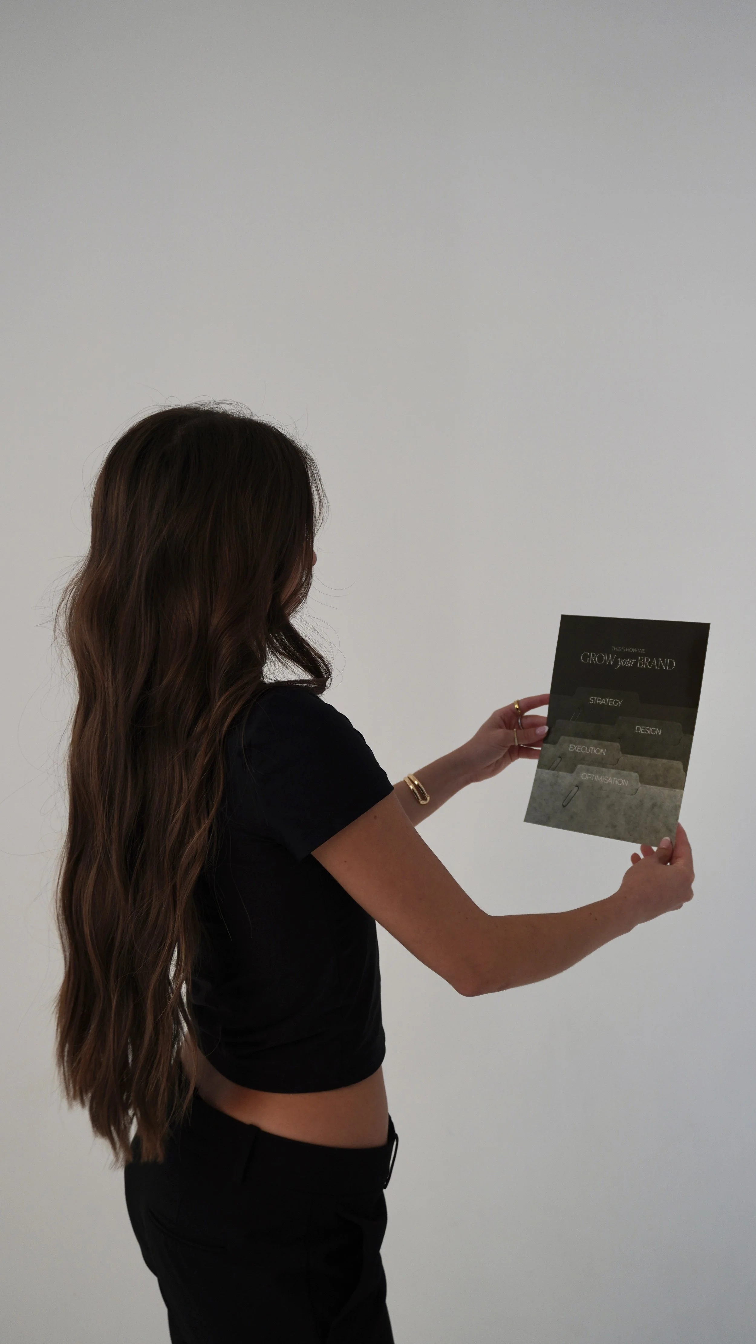 A woman with long wavy brown hair, wearing a black top and bracelets, is holding a brochure that reads "GROW your BRAND" and includes sections titled 'STRATEGY,' 'DESIGN,' 'EXECUTION,' and 'OPTIMISATION' against a plain white wall background.