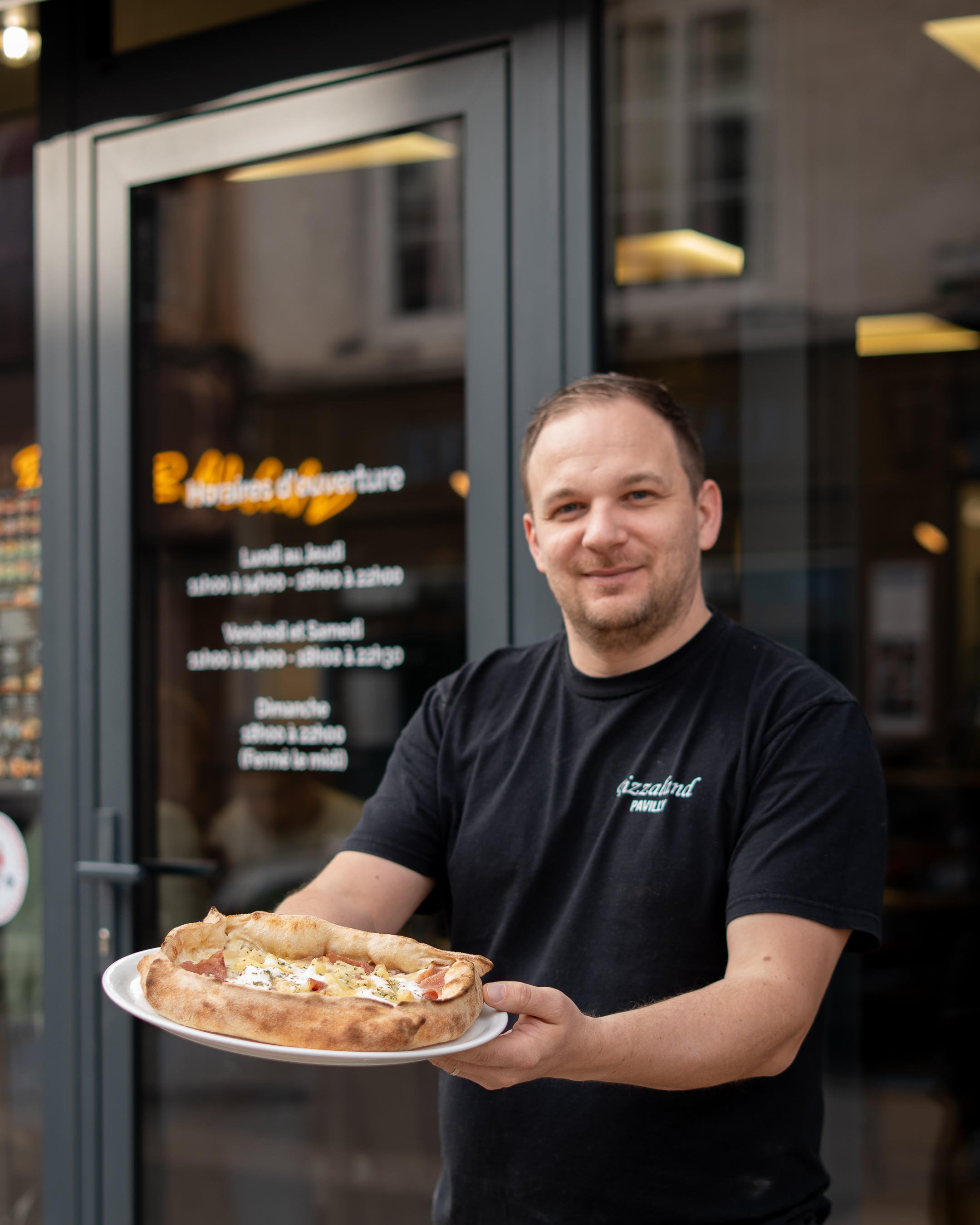 Un homme souriant tenant une pizza dans un restaurant ou pizzeria, avec une façade en verre et une enseigne lumineuse en arrière-plan.