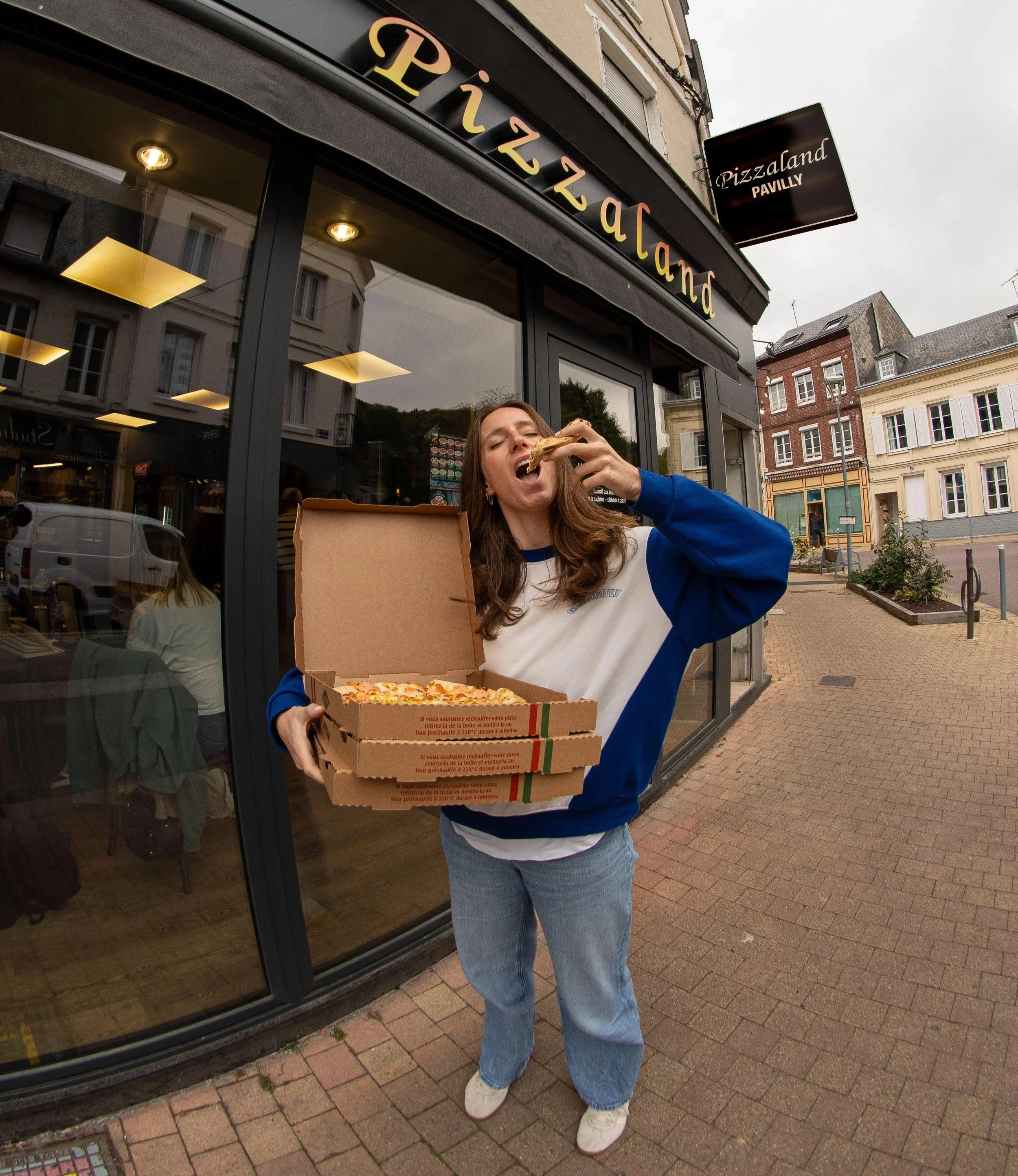 Une femme portant un sweat à capuche bleu et des jeans décontractés, tenant trois boîtes à pizza vides devant une pizzeria appelée 'Pizzaland'. Elle mange une part de pizza directement de la boîte. La devanture de la pizzeria a un grand panneau noir avec le nom 'Pizzaland' en lettres jaunes et un panneau suspendu indiquant 'Pizzaland PAVILL.'