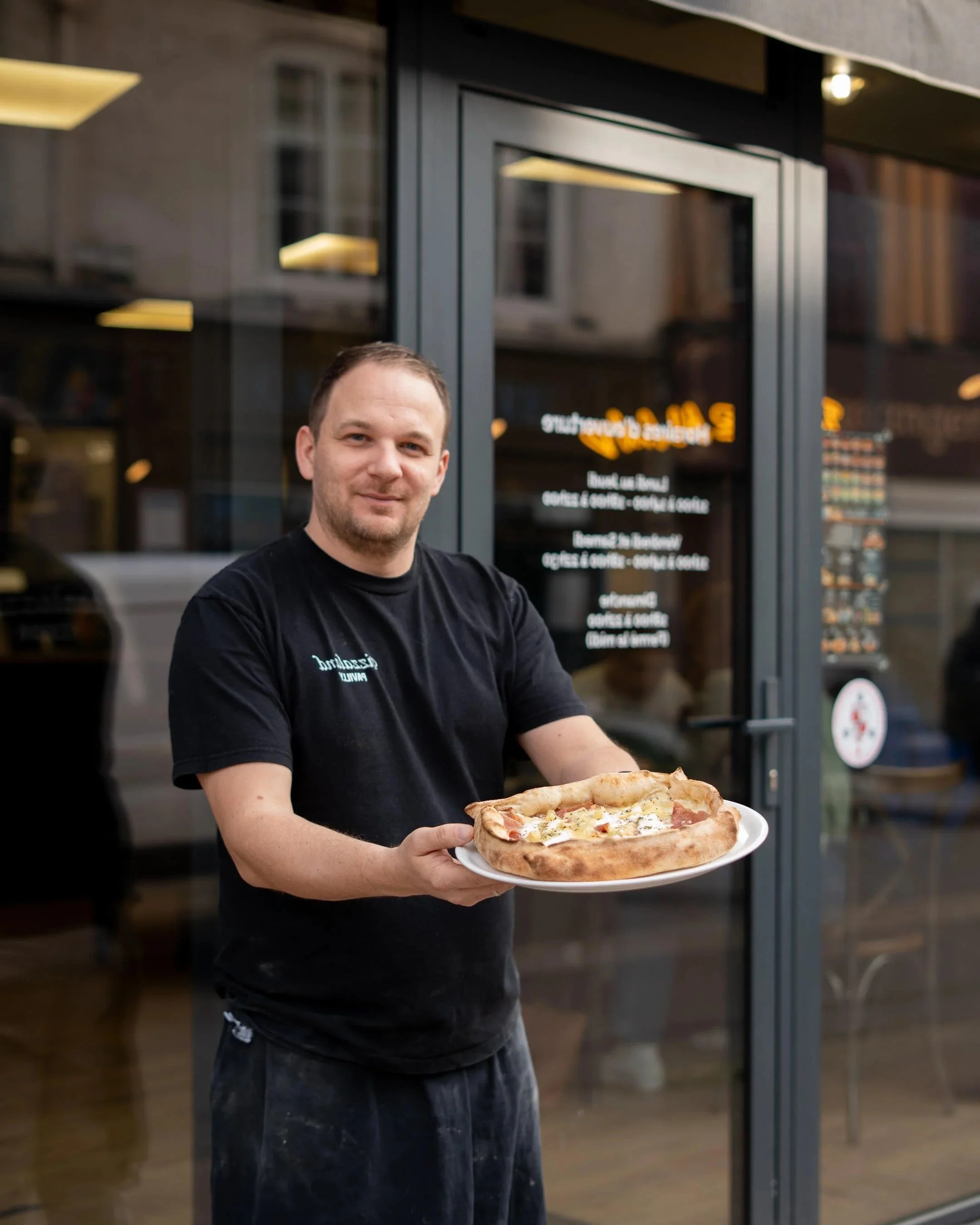 Un homme souriant, portant un t-shirt noir, tient une assiette avec une pizza dans une salle de restaurant ou de pizzeria.