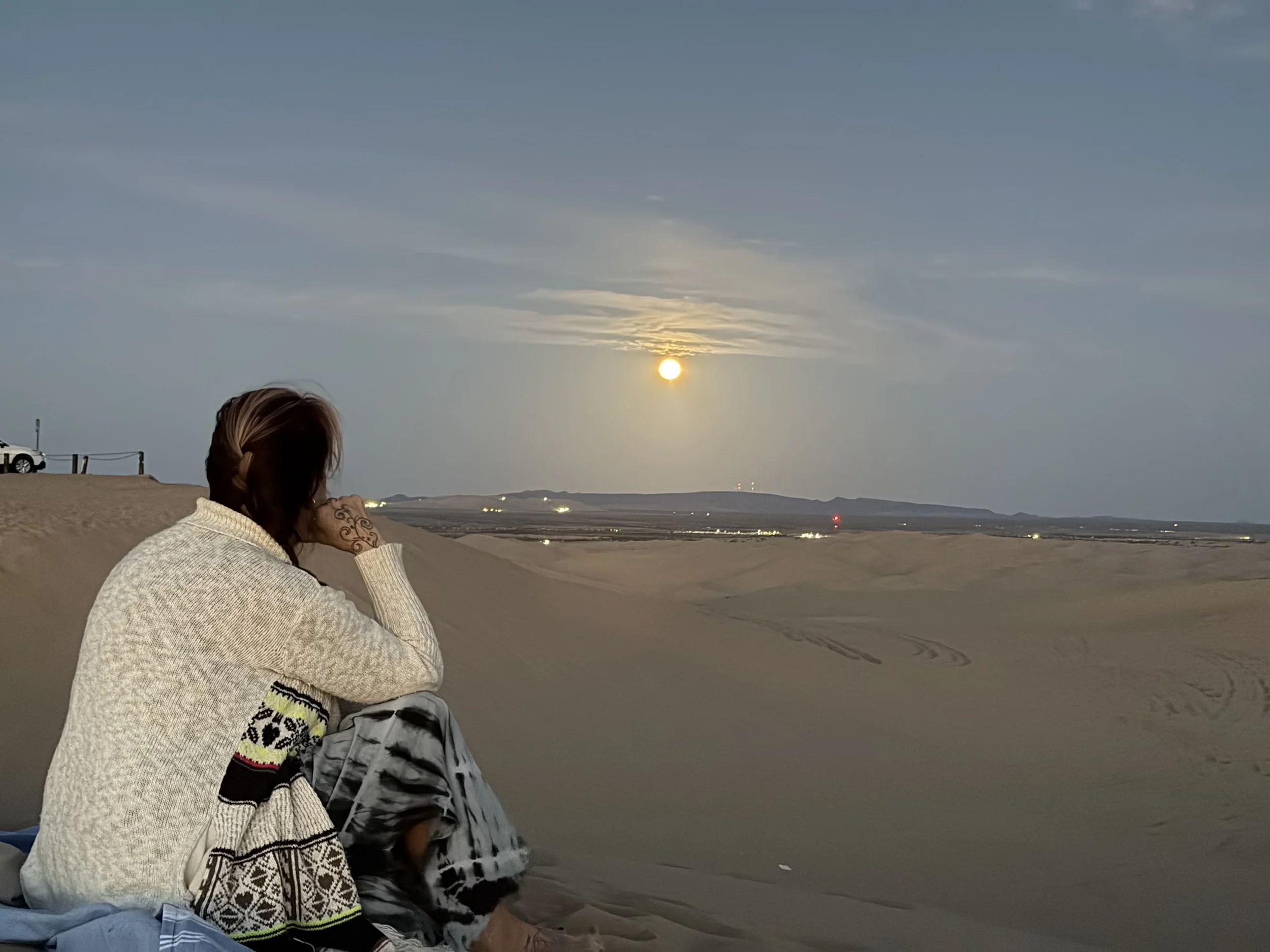 A person with long hair sitting on a sandy beach at sunset, wearing a cream sweater and patterned socks, looking towards the horizon. The sky is partly cloudy with the sun near the horizon, and there are distant hills and lights in the background.