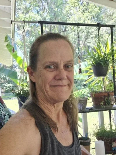 A woman taking a selfie on a porch or patio with green plants in the background.