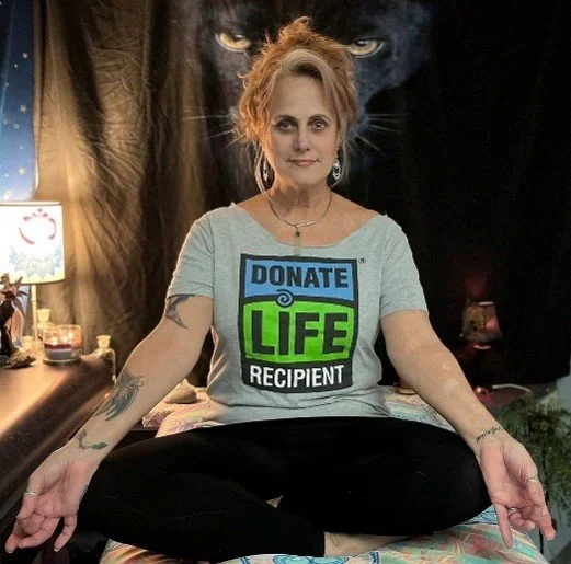 Woman sitting cross-legged on bed doing yoga, wearing a t-shirt that says 'Donate Life Recipient', with a dark wolf face in the background and a small lamp on a nearby dresser.