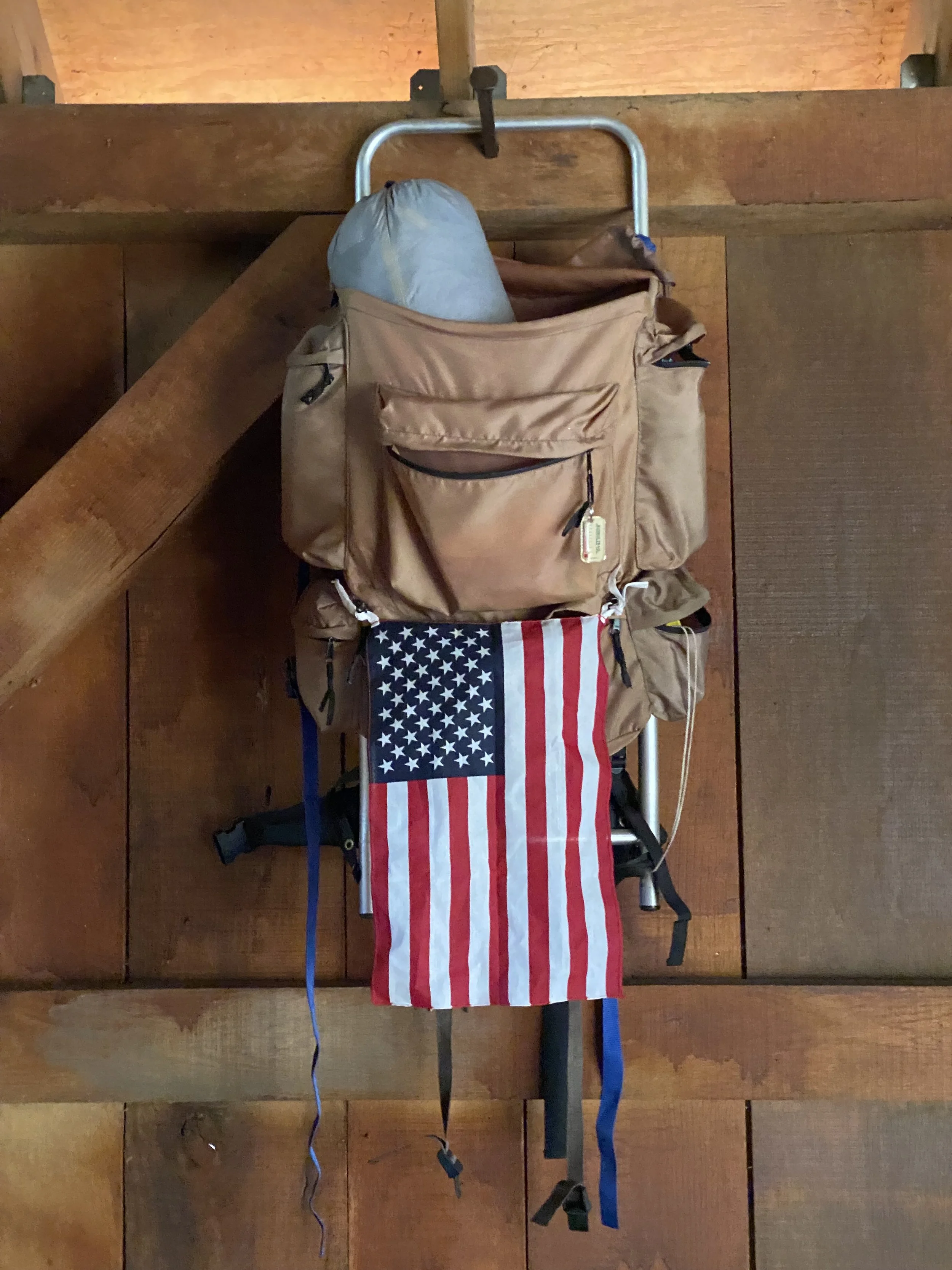 A backpack with an American flag draped on it, hanging on a metal frame hanger on a wooden wall.