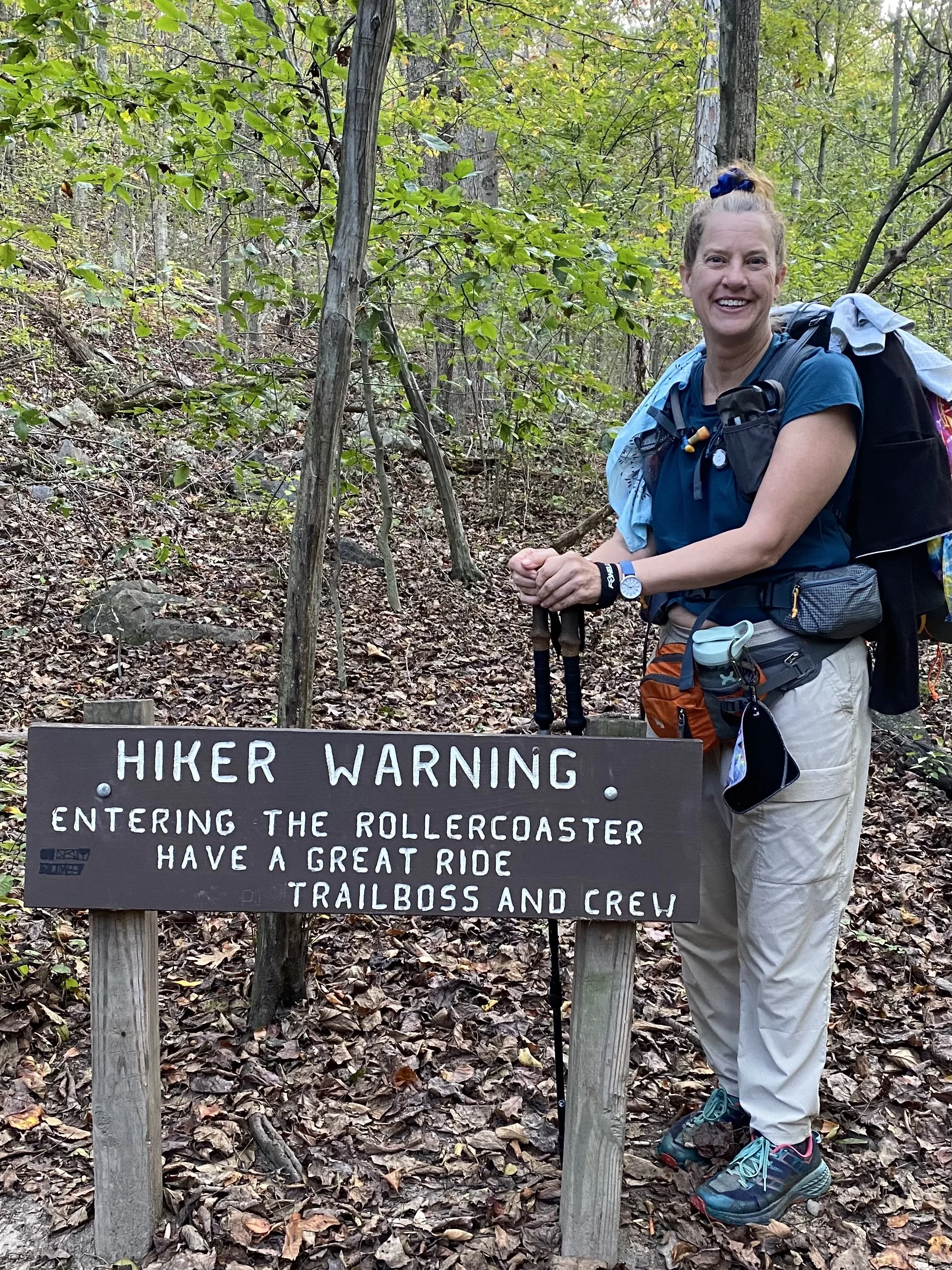 A woman standing next to a sign that reads, '.Hiker Warning. Entering the rollercoaster have a great ride to trailboss and crew.' She is in a wooded area, smiling, wearing a blue shirt, khaki pants, hiking shoes, and carrying a backpack with gear attached.