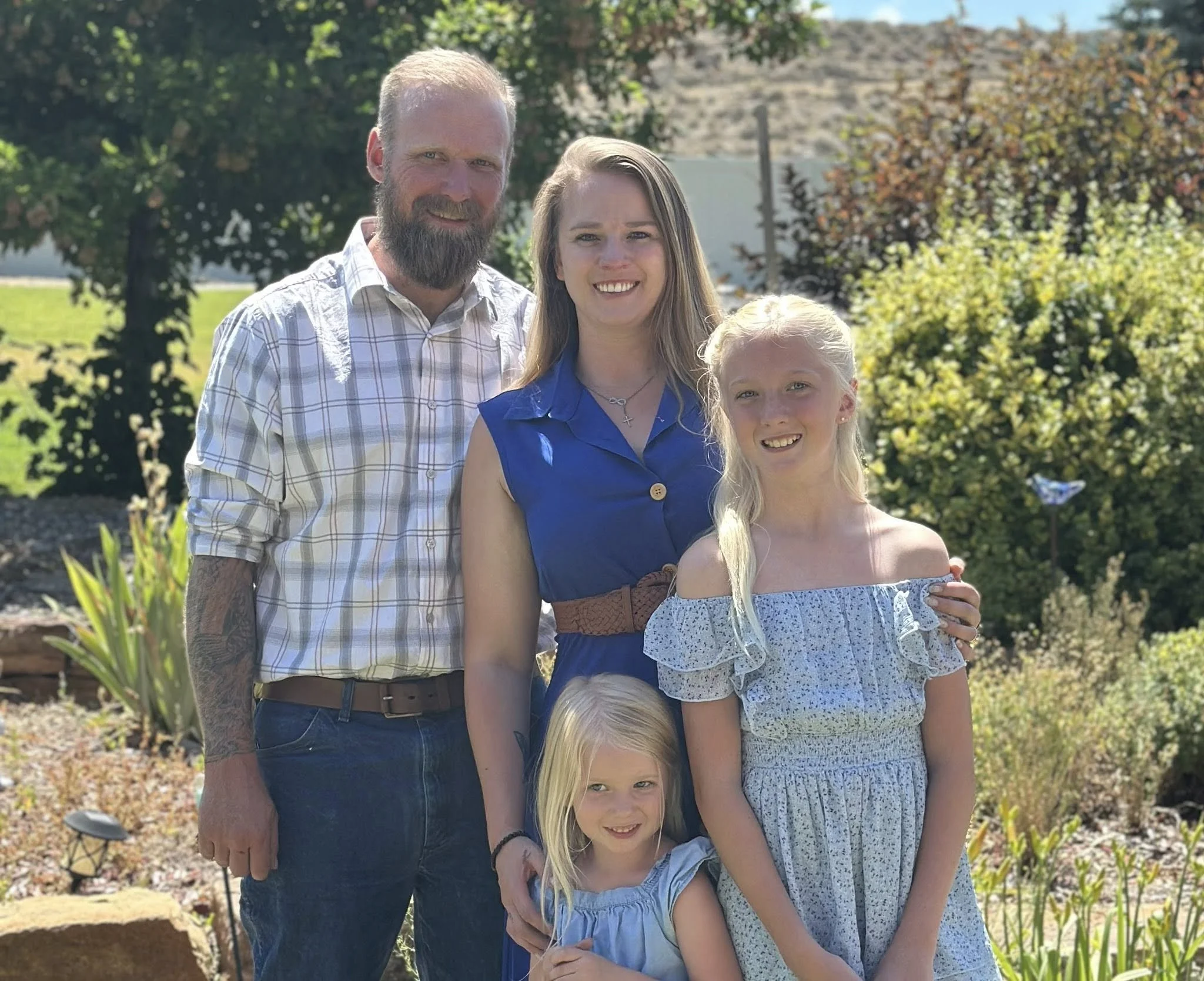 A family of five standing outdoors in a garden during daytime. The family includes two adults and three children. They are smiling at the camera with trees and greenery in the background.