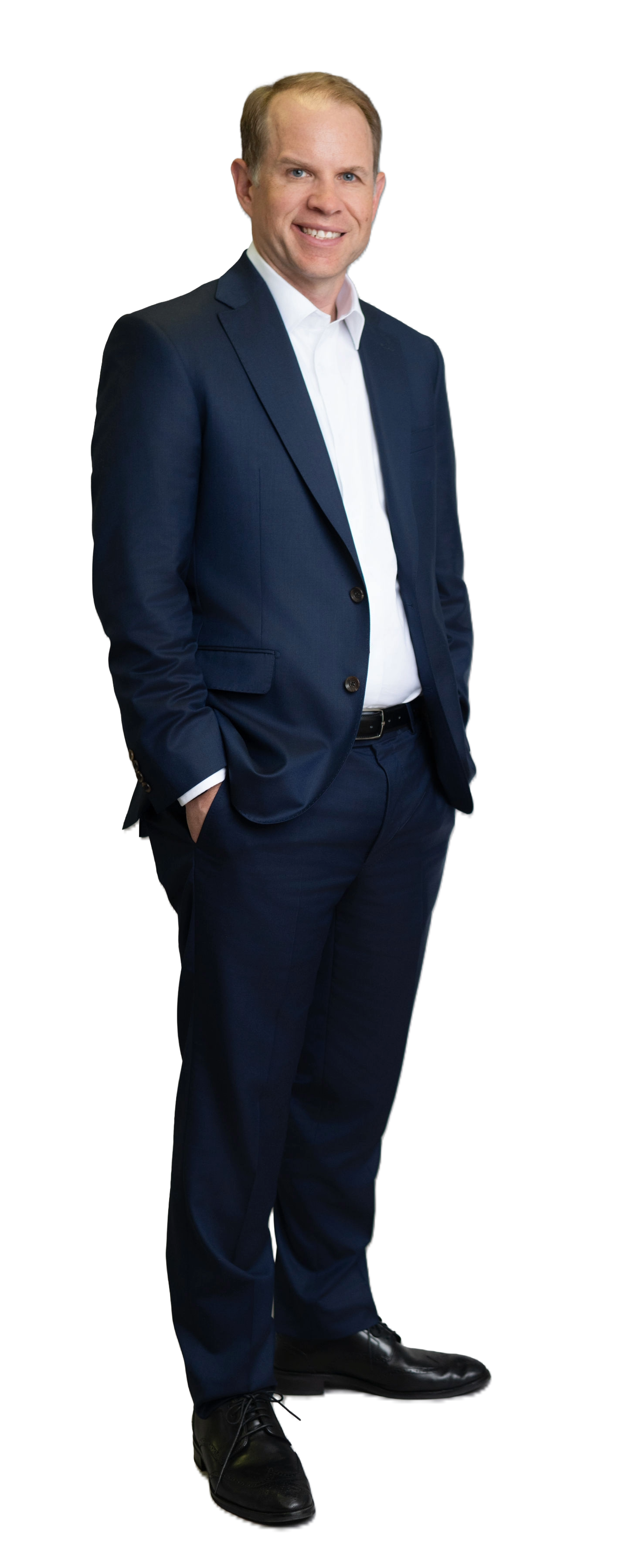 A man in a dark blue suit, white shirt, and black shoes standing with hands in pockets and smiling.