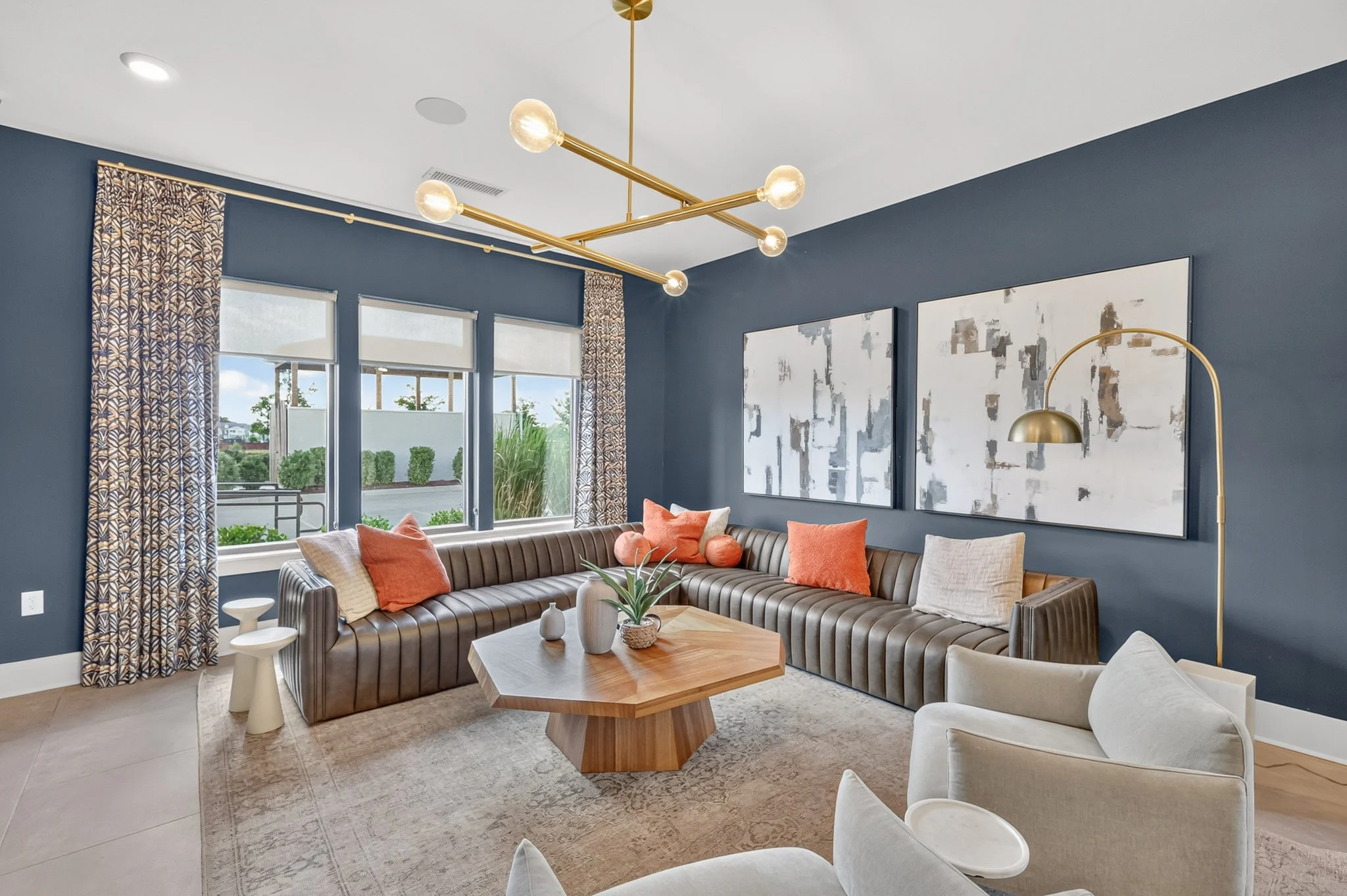 Living room with blue walls, large windows with patterned curtains, modern gold chandelier, abstract artwork, beige and white chairs, and a wooden coffee table with plants.