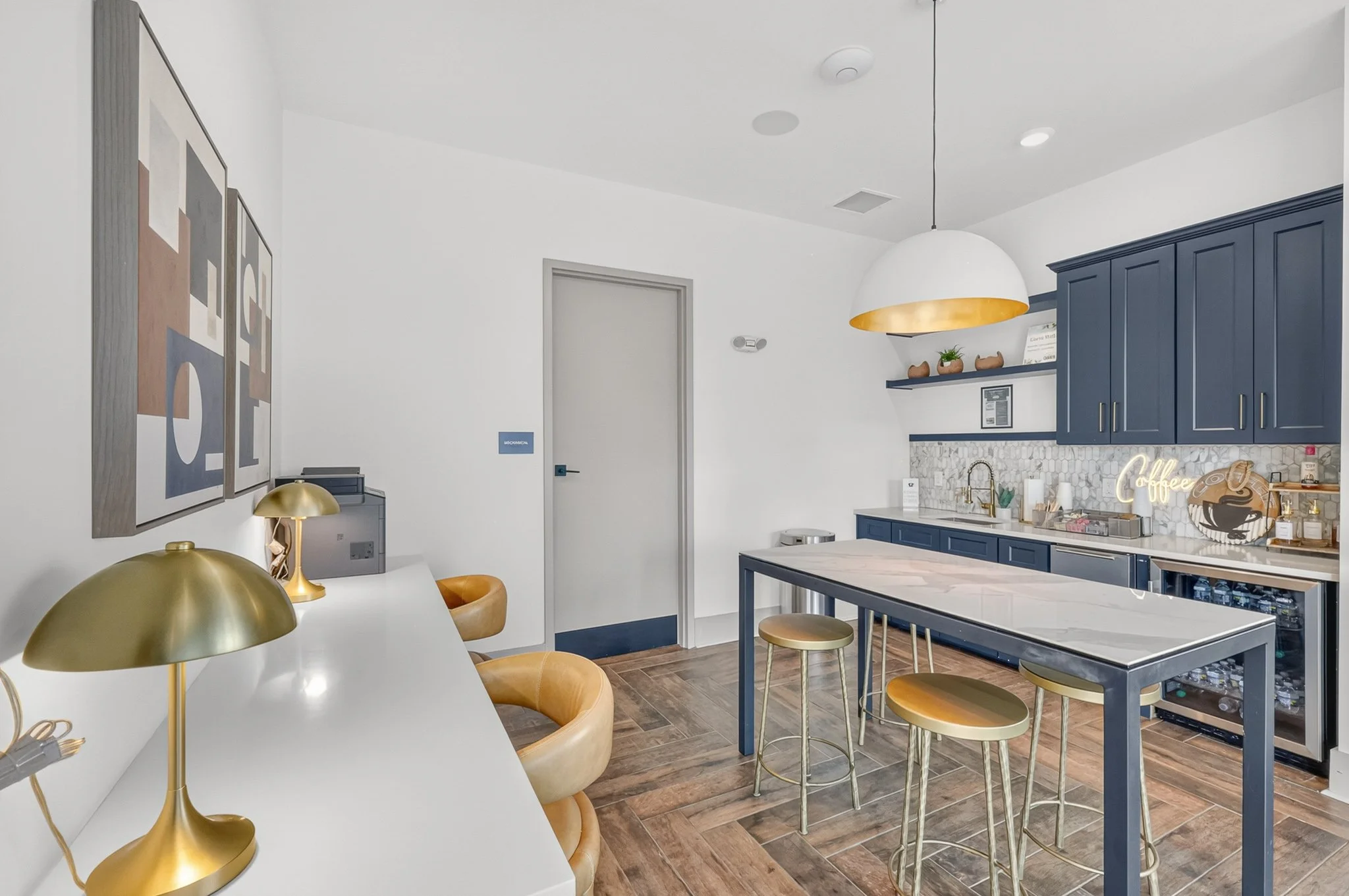 A modern kitchen with navy blue cabinets, a marble countertop, wooden bar stools, and a hanging white and gold pendant light. Decor includes small plants, a neon coffee sign, and decorative items.