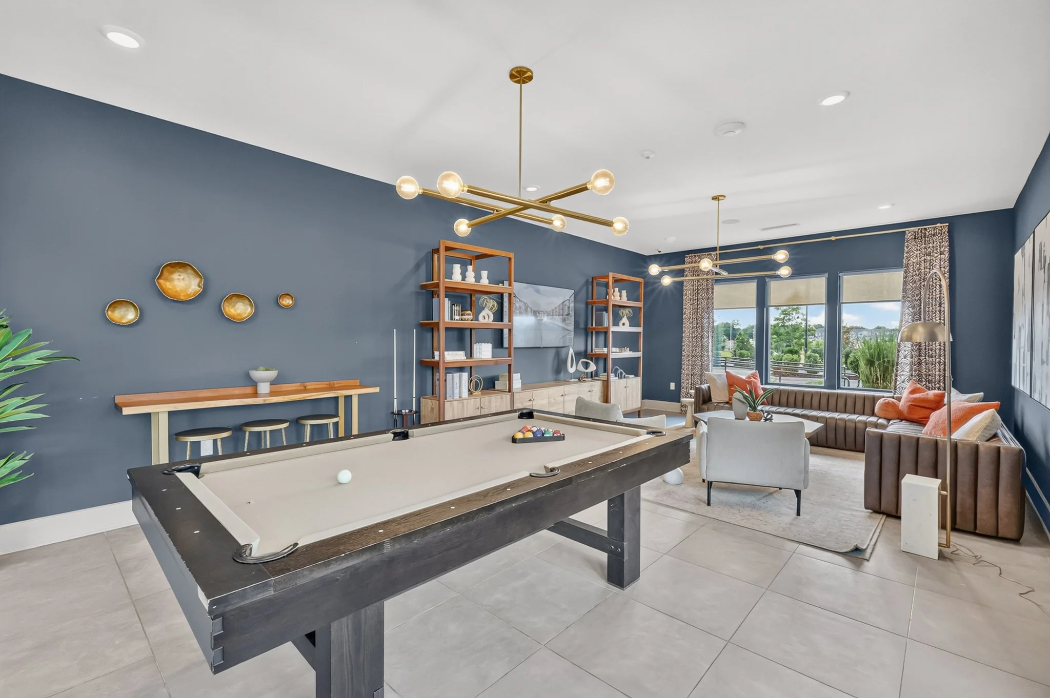 Living room with a pool table, blue accent wall, modern chandelier, wooden shelves, a tan sectional sofa, and large windows with patterned curtains.