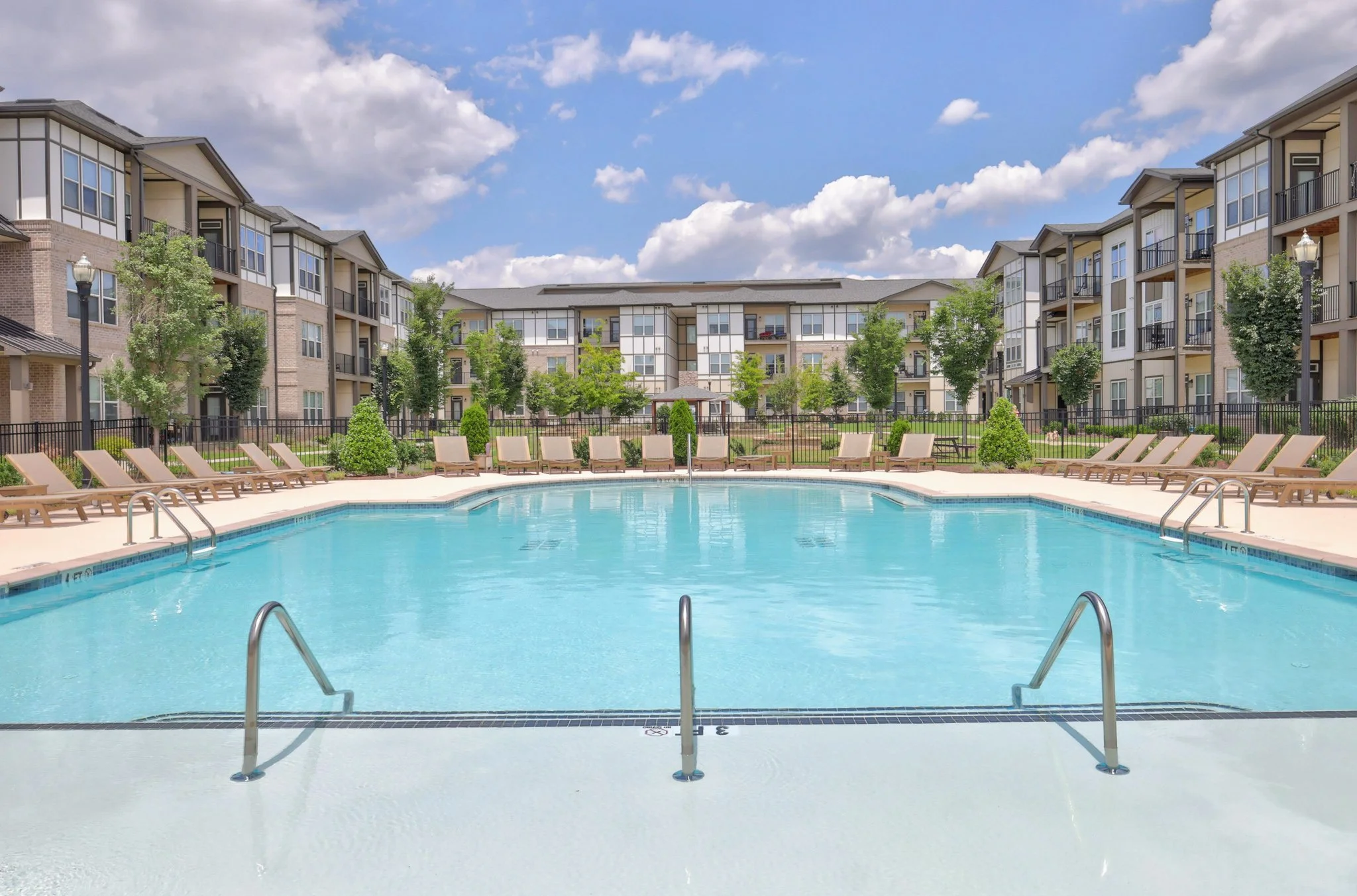 An outdoor swimming pool with clear blue water, surrounded by lounge chairs, trees, and a fence, with modern apartment buildings in the background under a partly cloudy sky.