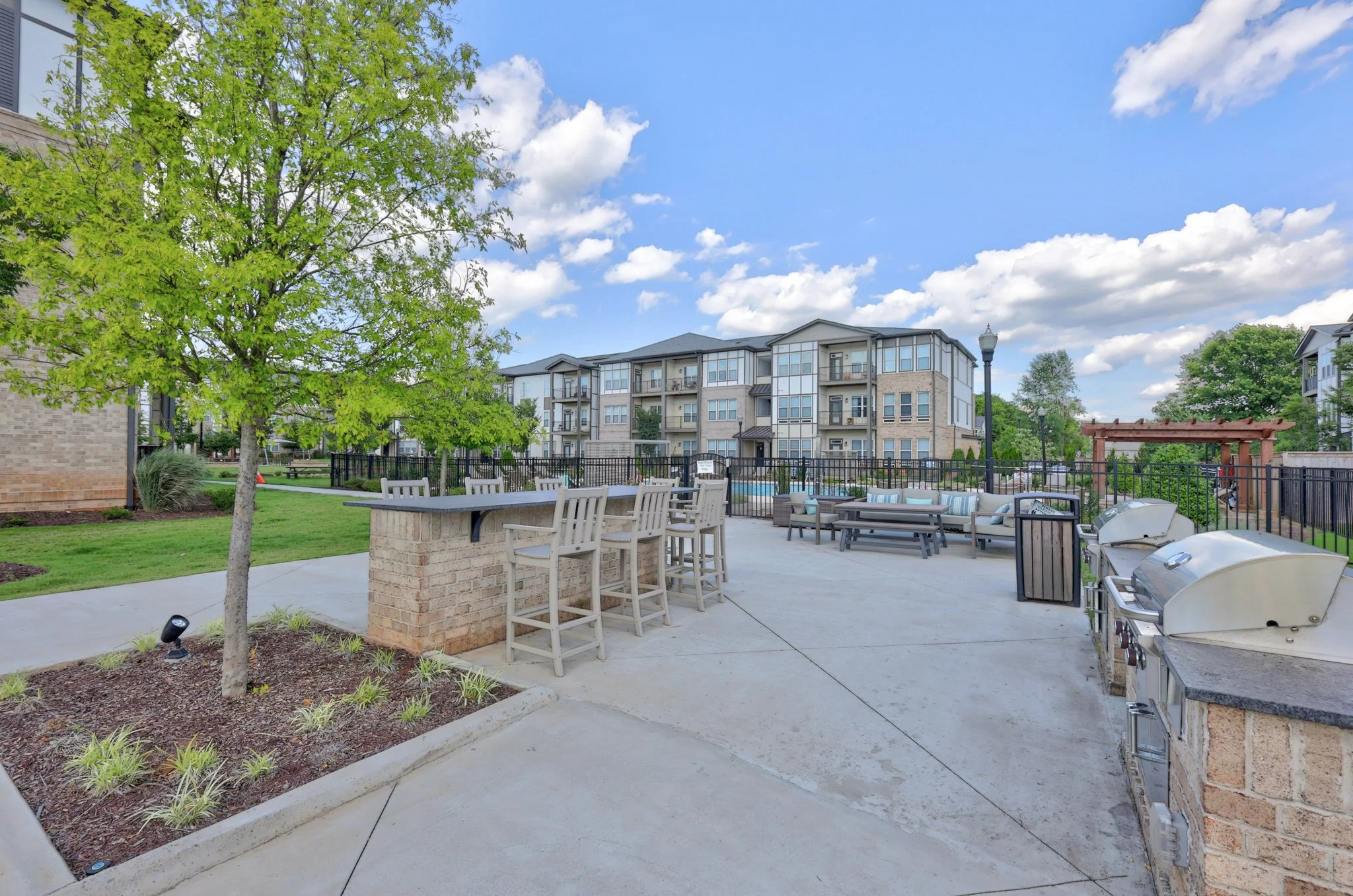 An outdoor patio area with a green lawn, a large tree, and chairs. In the background, there are multi-story apartment buildings and a blue sky with scattered clouds.