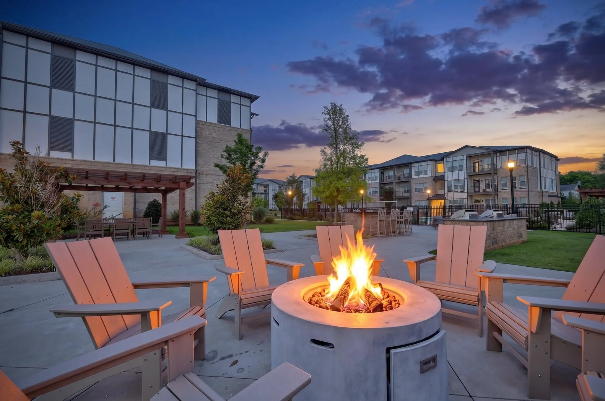 Exterior courtyard of an apartment complex at dusk with lit fire pit, surrounded by chairs, trees, and modern multi-story buildings under a partly cloudy sky.